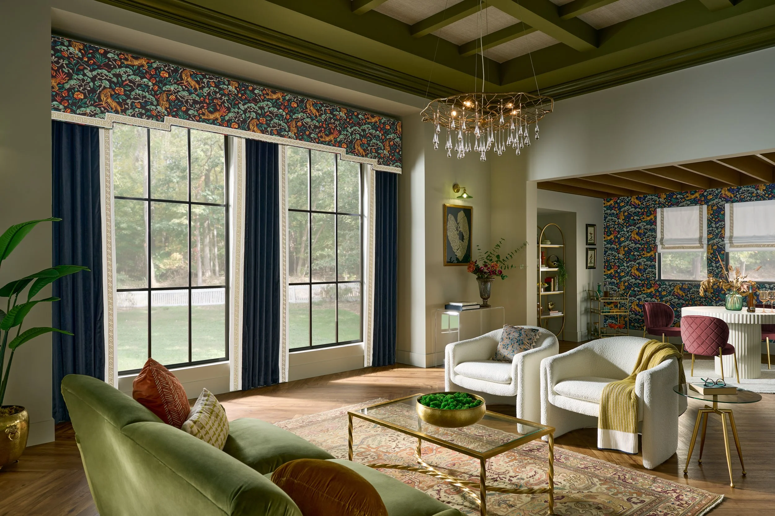 Spacious living room with large windows, blue curtains, and a colorful floral and tiger wallpaper accent wall. Furniture includes a green velvet sofa, white armchairs, a gold coffee table, and a velvet pink dining chair. Decor features a modern chandelier with hanging crystals, framed artwork, flowers, and bookshelf.