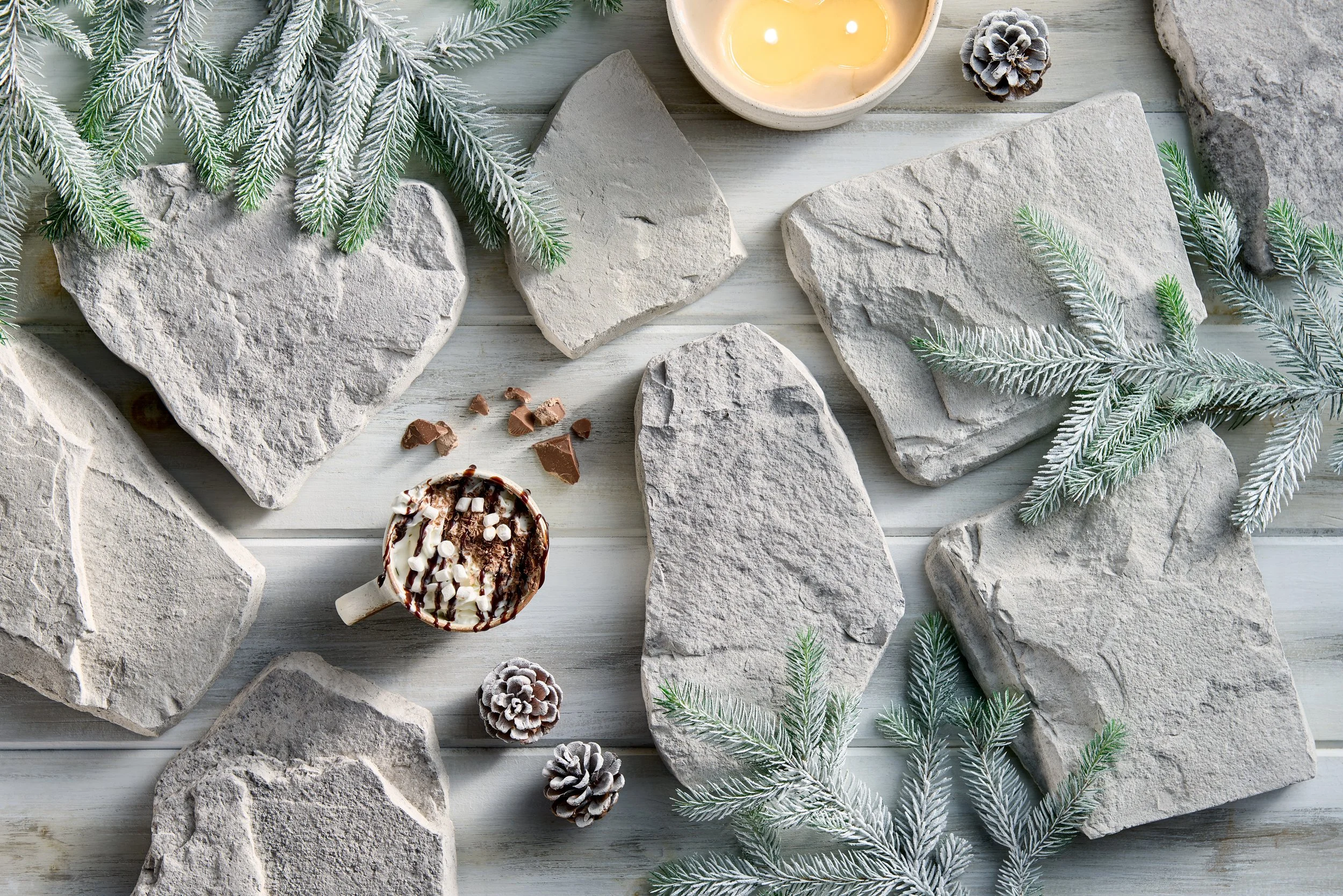 Flat lay of gray rocks, pine branches with snow, pinecones, a bowl of melted wax, and a cup of hot chocolate topped with whipped cream and sprinkles on a white wooden surface.