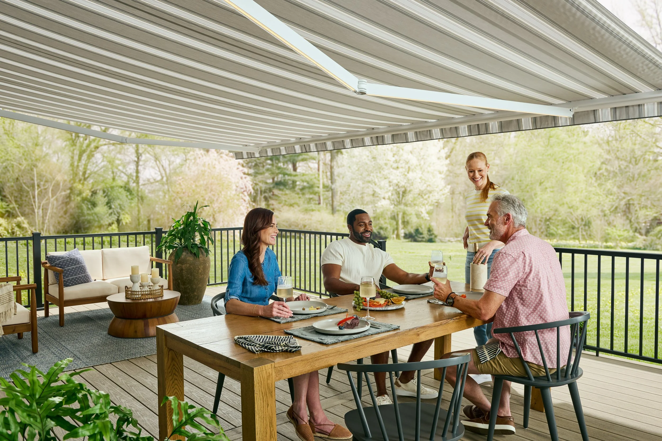 A group of people enjoying a meal together on a covered outdoor patio. They are seated around a wooden table with food and drinks, smiling and engaging in conversation. The patio has a metal railing and is surrounded by green trees and grass.