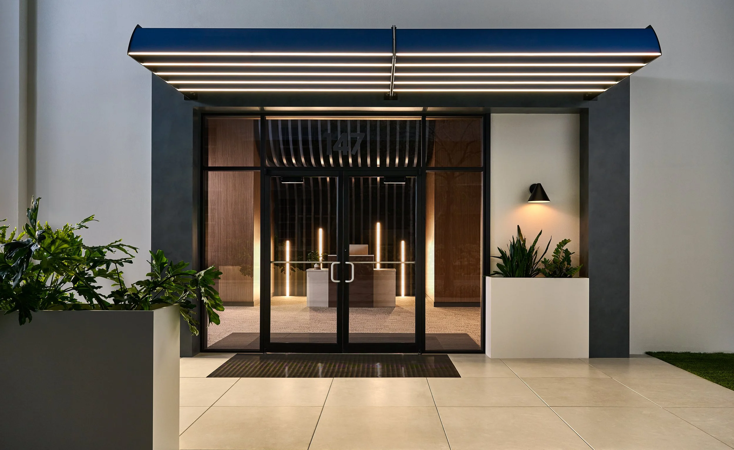 Modern glass doorway with black frame, illuminated interior, surrounded by white and grey walls, with potted green plants outside on either side.