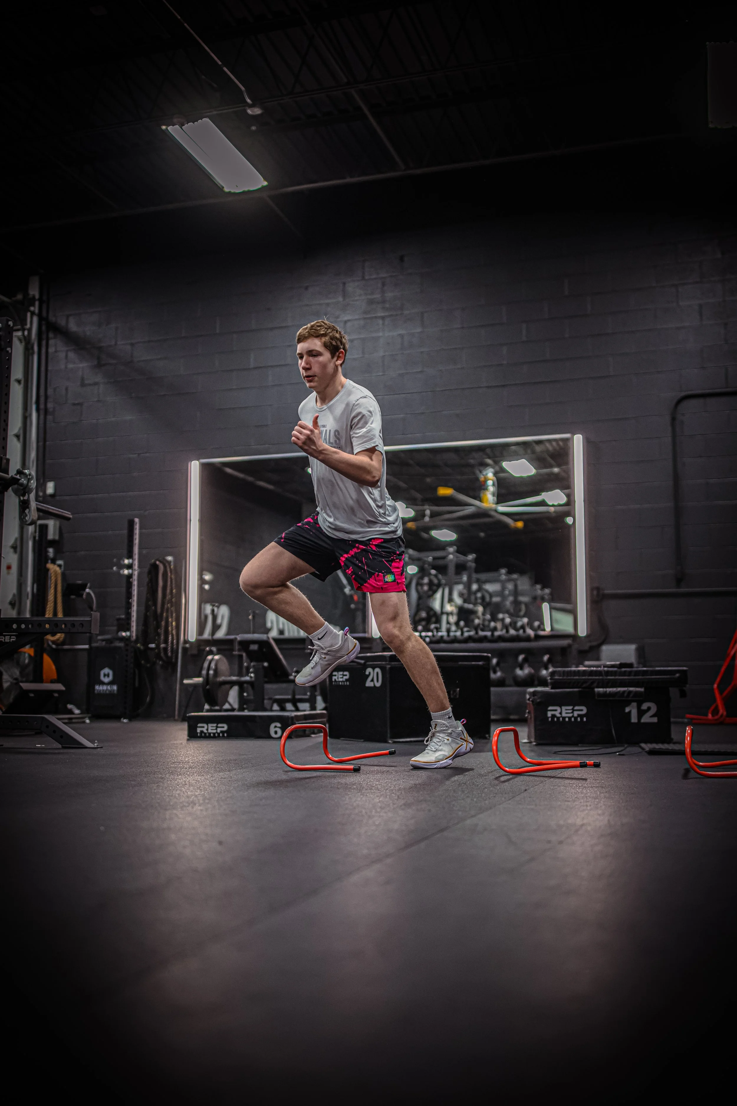 A young man wearing a gray t-shirt and black shorts with pink accents performs a box jump over agility hurdles inside a gym with black walls and fitness equipment in the background.