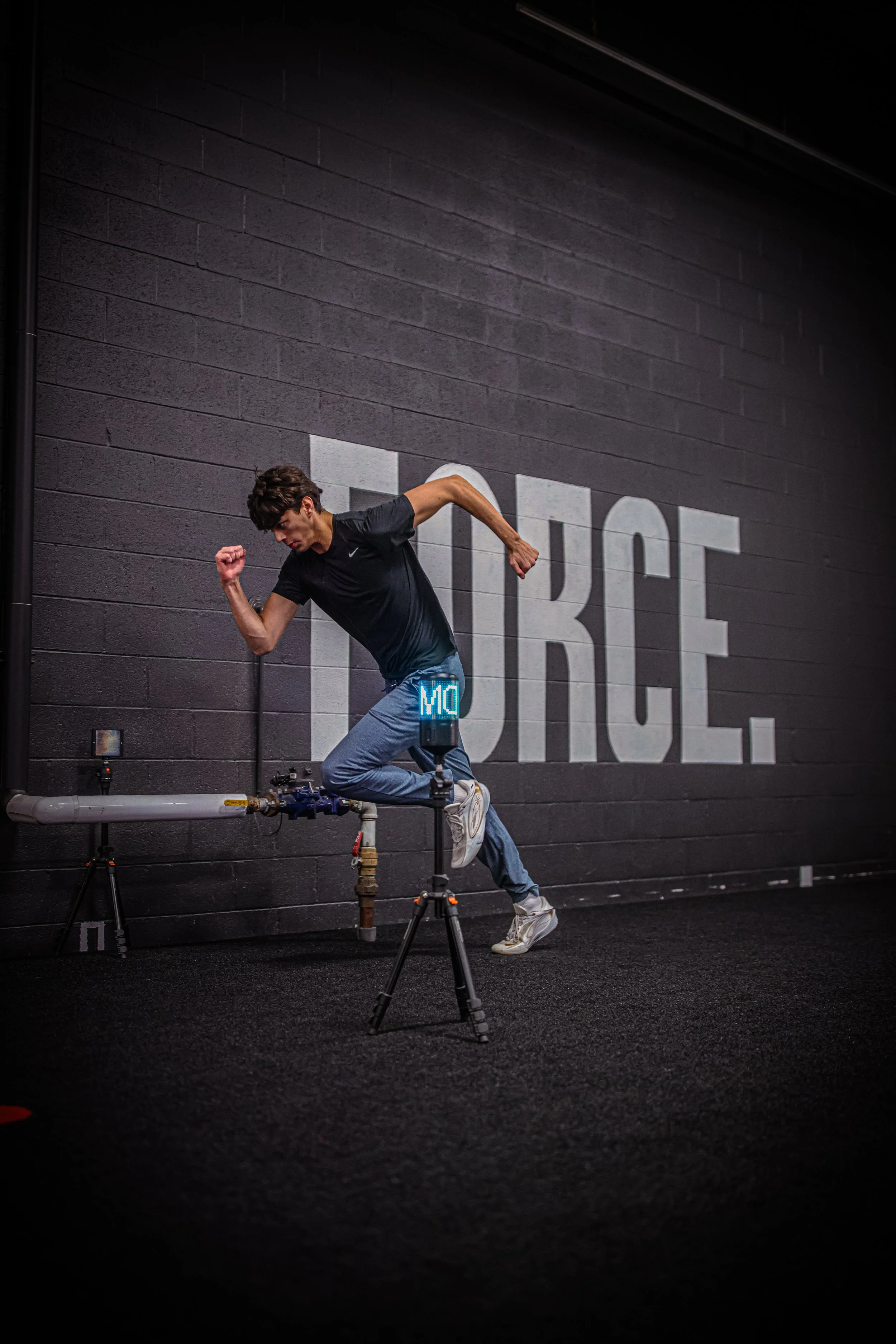 A young man running indoors on a treadmill that is built into the wall, with a dark brick wall and the words 'RISE' visible in large letters behind him.