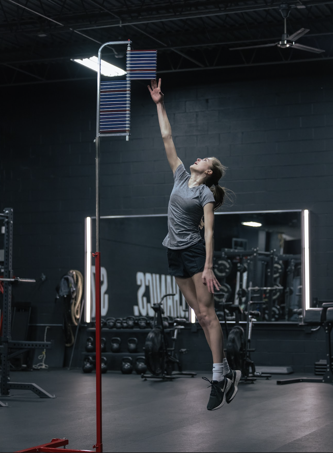A woman in athletic clothing performing a high jump on a fitness tracker in a gym with black walls, gym equipment, and a large mirror.