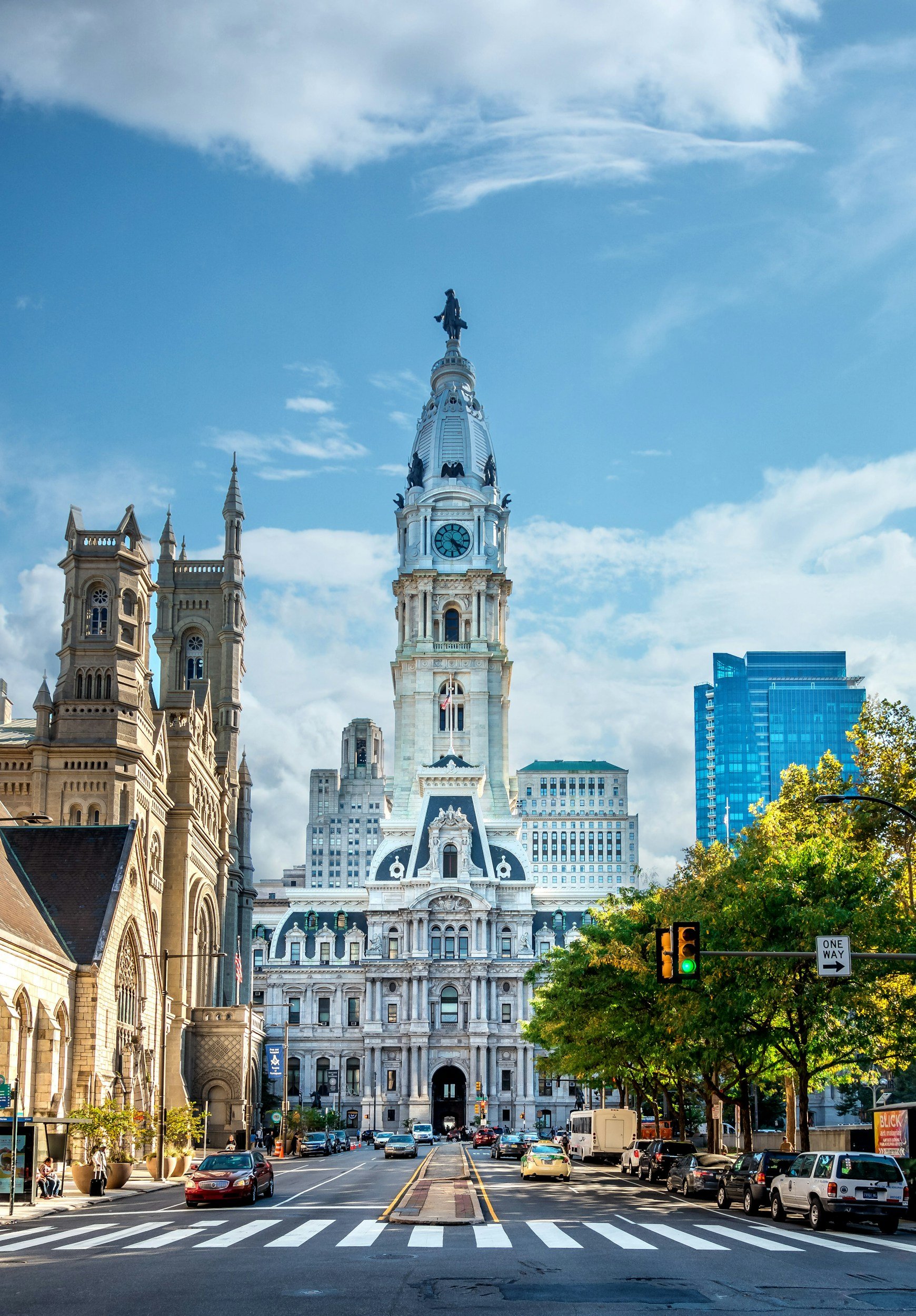 Philadelphia City Hall with a clock tower and statue on top, surrounded by city buildings, trees, and cars on the street.