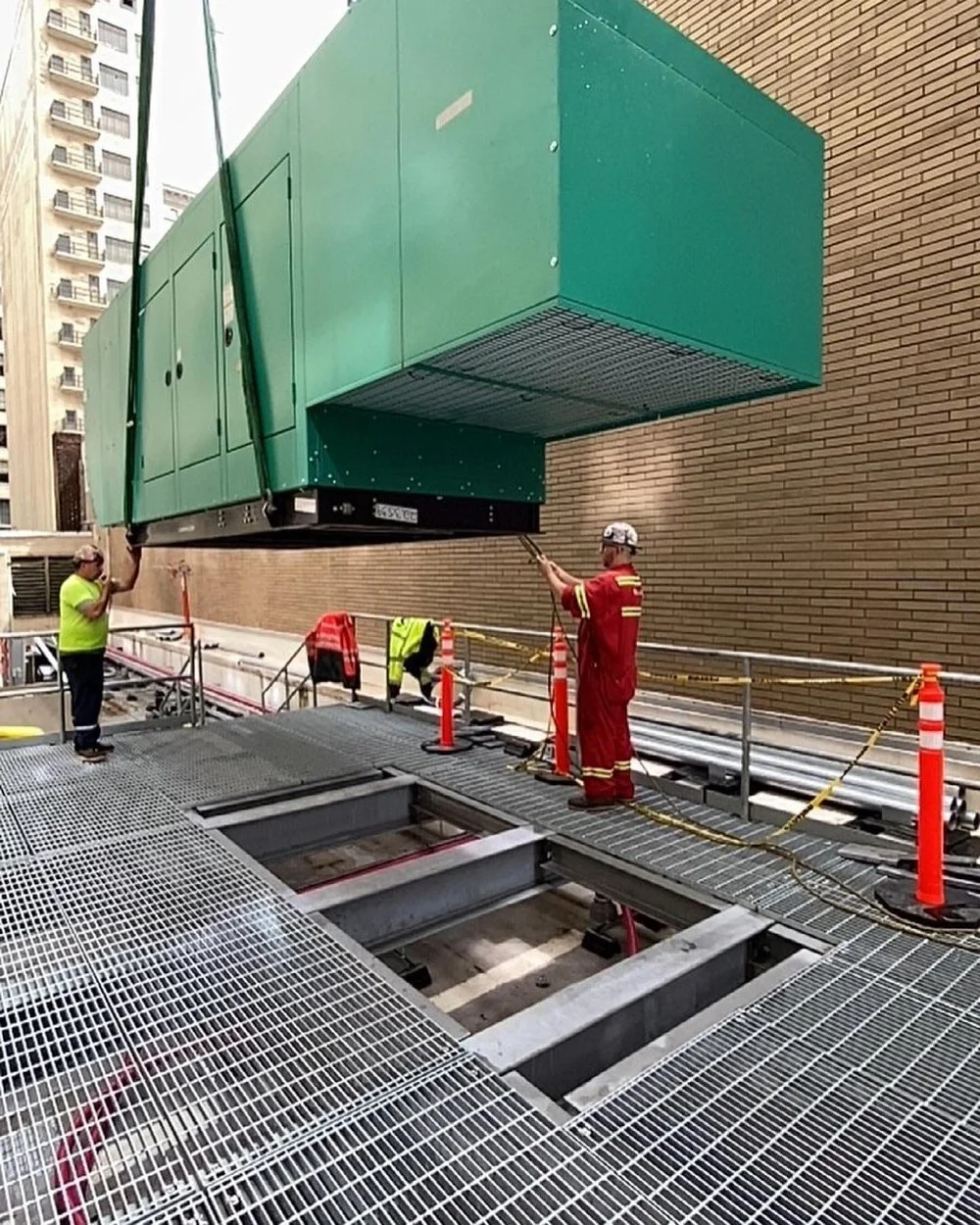 Two workers noise testing or inspecting a large green HVAC unit suspended above an industrial rooftop by metal cables, with metal grated flooring in the foreground and a brick building wall in the background.