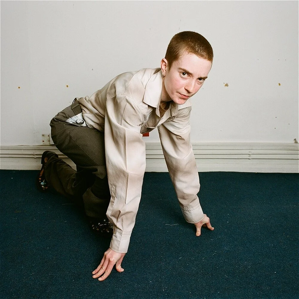 Slim white person stands with enormous arms, posing to show off their muscles. They are wearing a brown t-shirt, grey trousers with white paint smeared over the crotch and shoes with shiny stickers stuck to them. The sky is dark but the person, tree