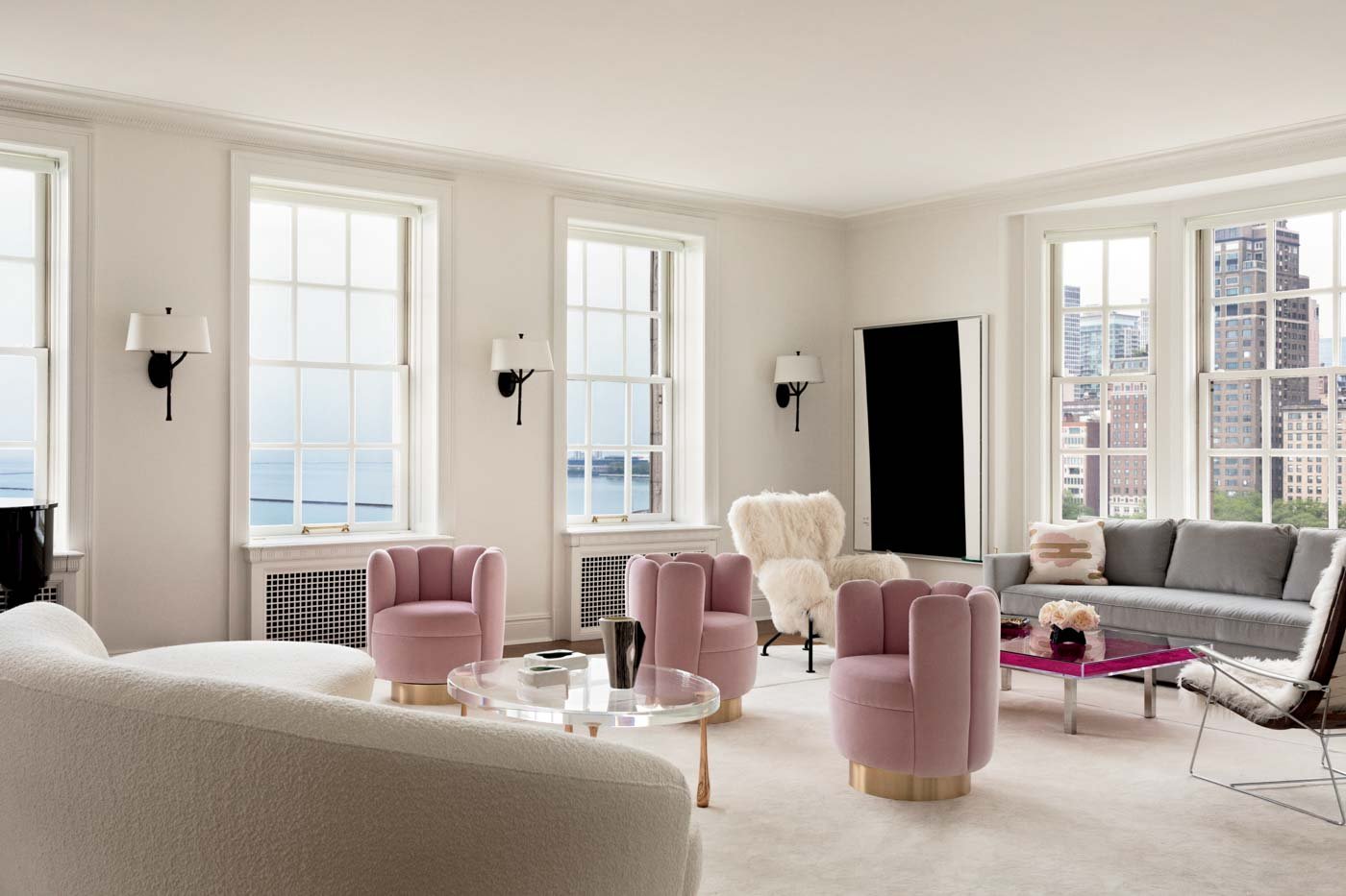 Modern living room with white walls, large windows, pink armchairs, a white sofa, a fluffy white armchair, black wall sconces, a pink coffee table with flowers, and a cityscape view.