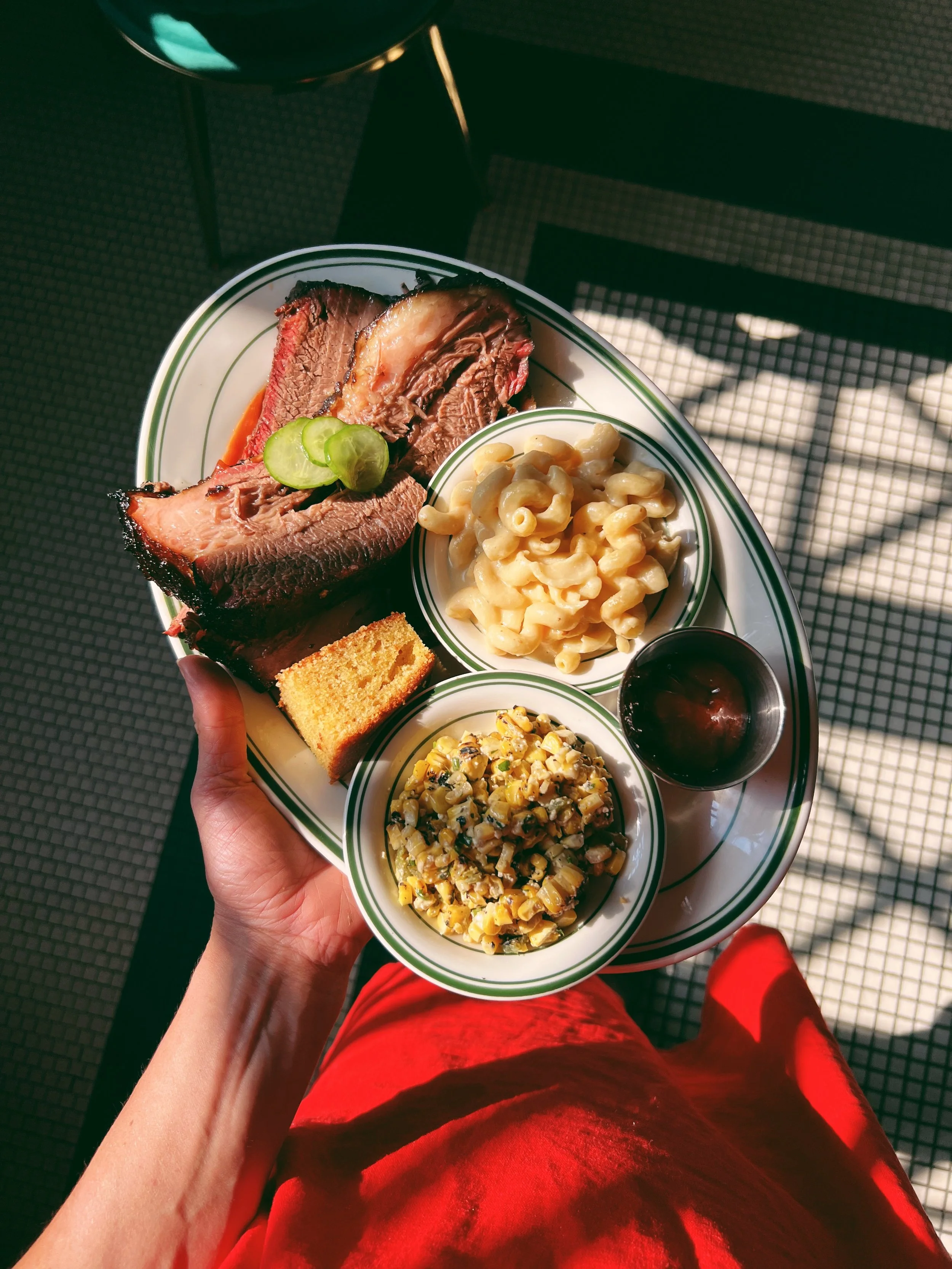 A plate with barbeque ribs garnished with pickles, a serving of macaroni and cheese, a small bowl of baked beans, a side of macaroni salad, a piece of cornbread, and a cup of barbecue sauce, held in a person's hand.