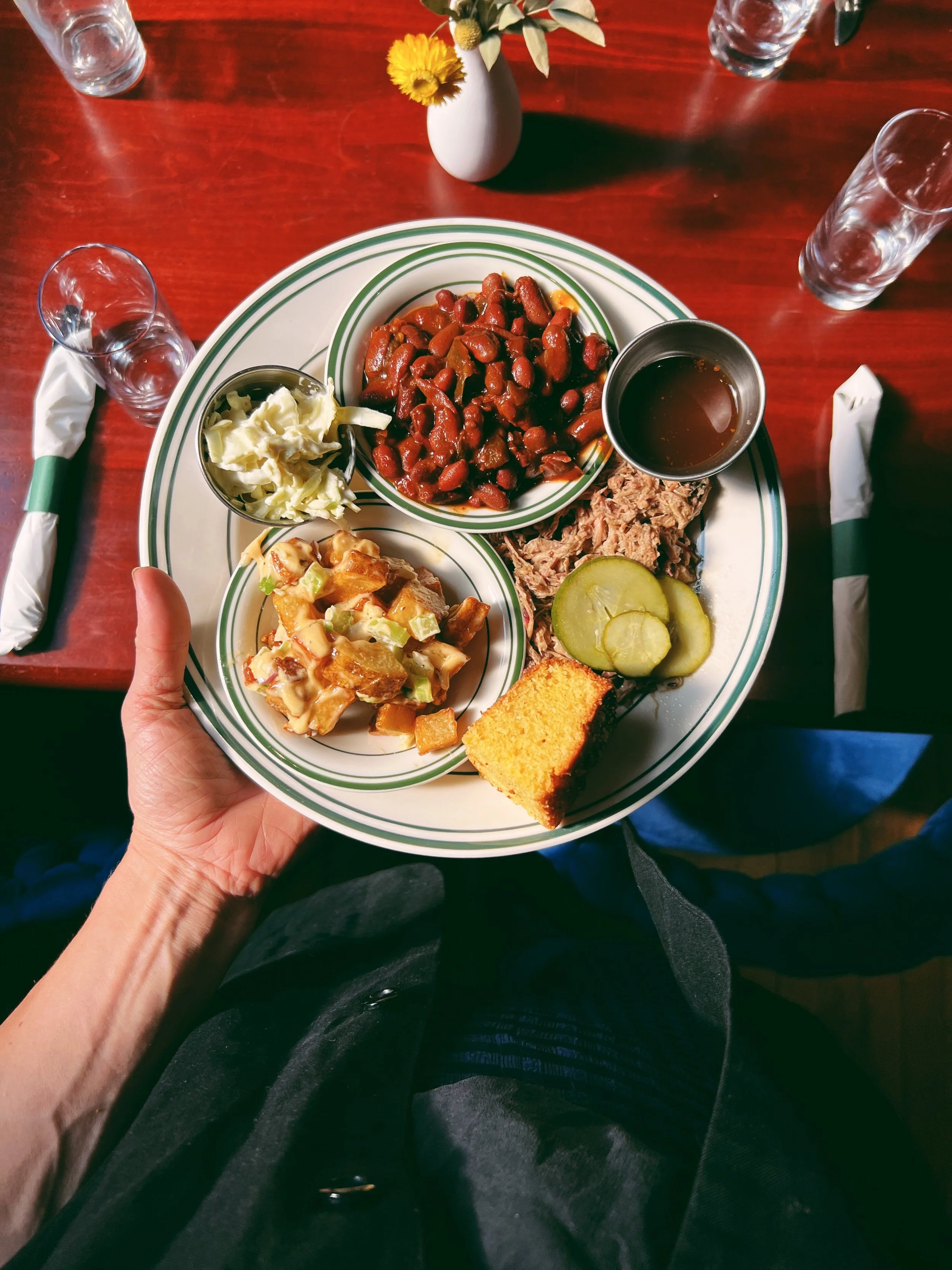 A person holding a plate with baked beans, coleslaw, BBQ pulled pork, pickles, cornbread, and a cup of barbecue sauce at a restaurant table with water glasses and a small flower vase.
