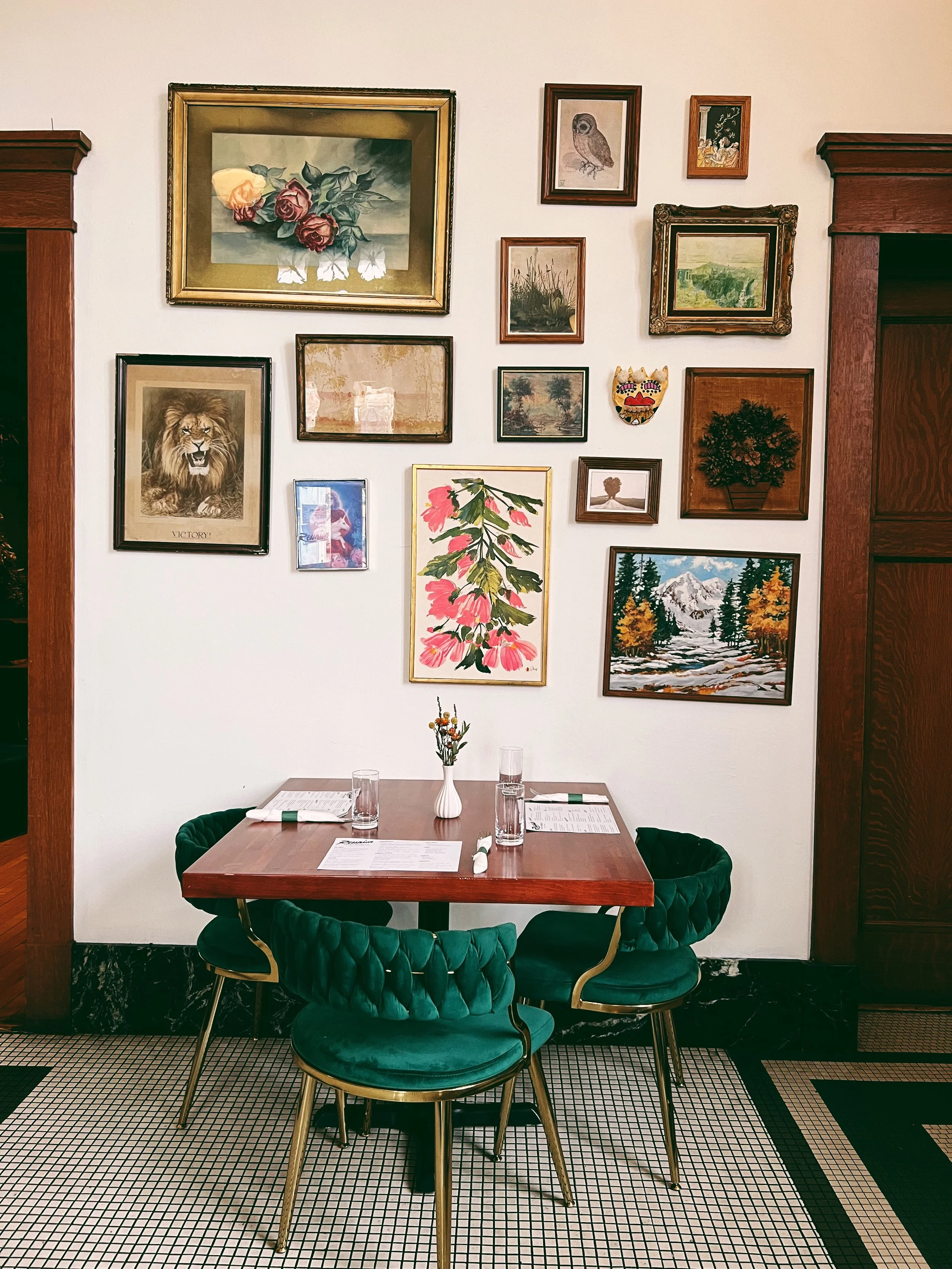 A small wooden table with four green velvet chairs, set with glasses, menus, and a small vase with flowers, against a white wall decorated with various framed artworks including paintings and prints of animals, landscapes, and floral designs, in a co