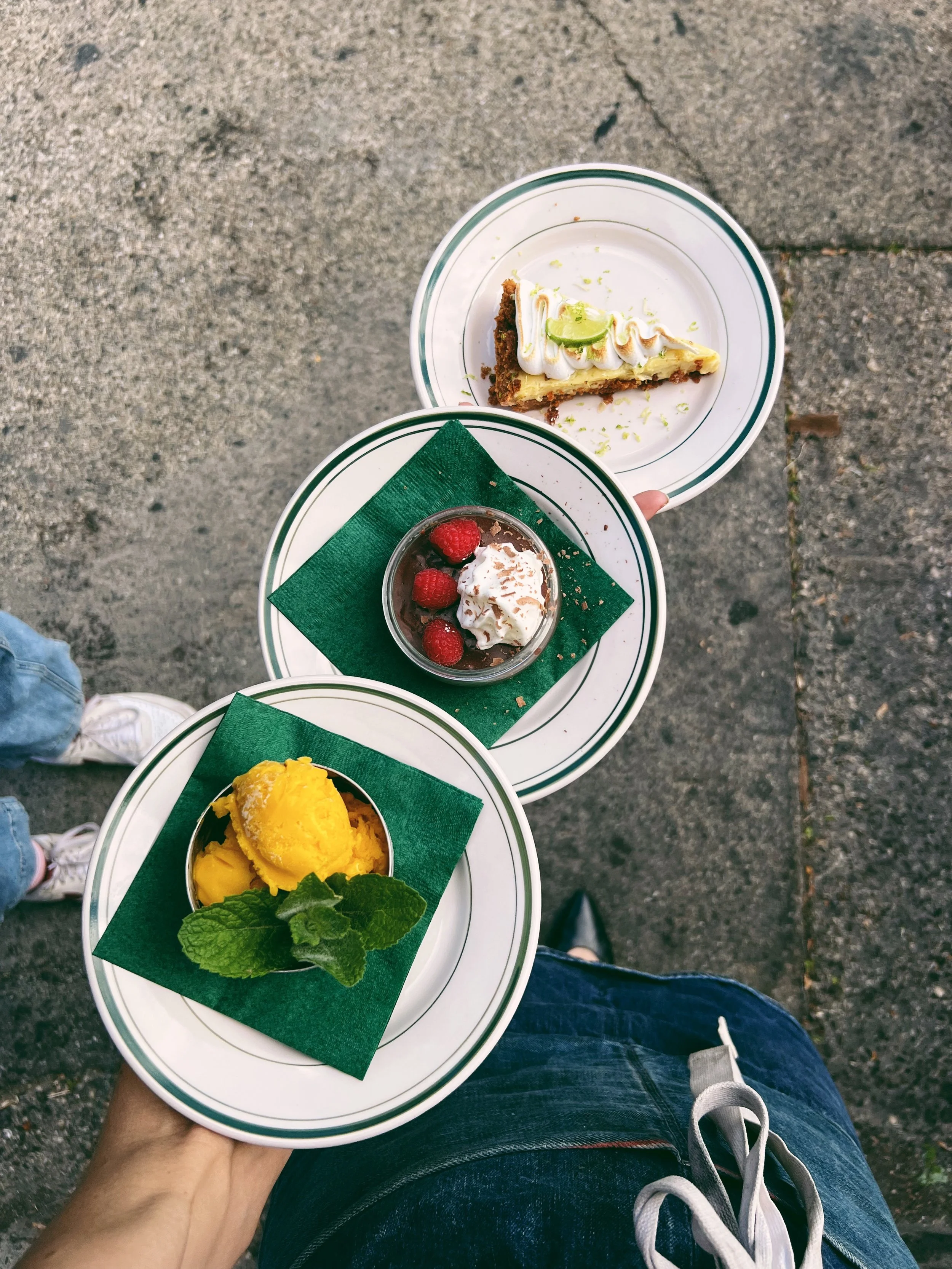 Three plates with desserts: a slice of lime-topped cake, a raspberry chocolate mousse, and a scoop of yellow ice cream garnished with mint leaves, held by a person outdoors on a gray pavement.