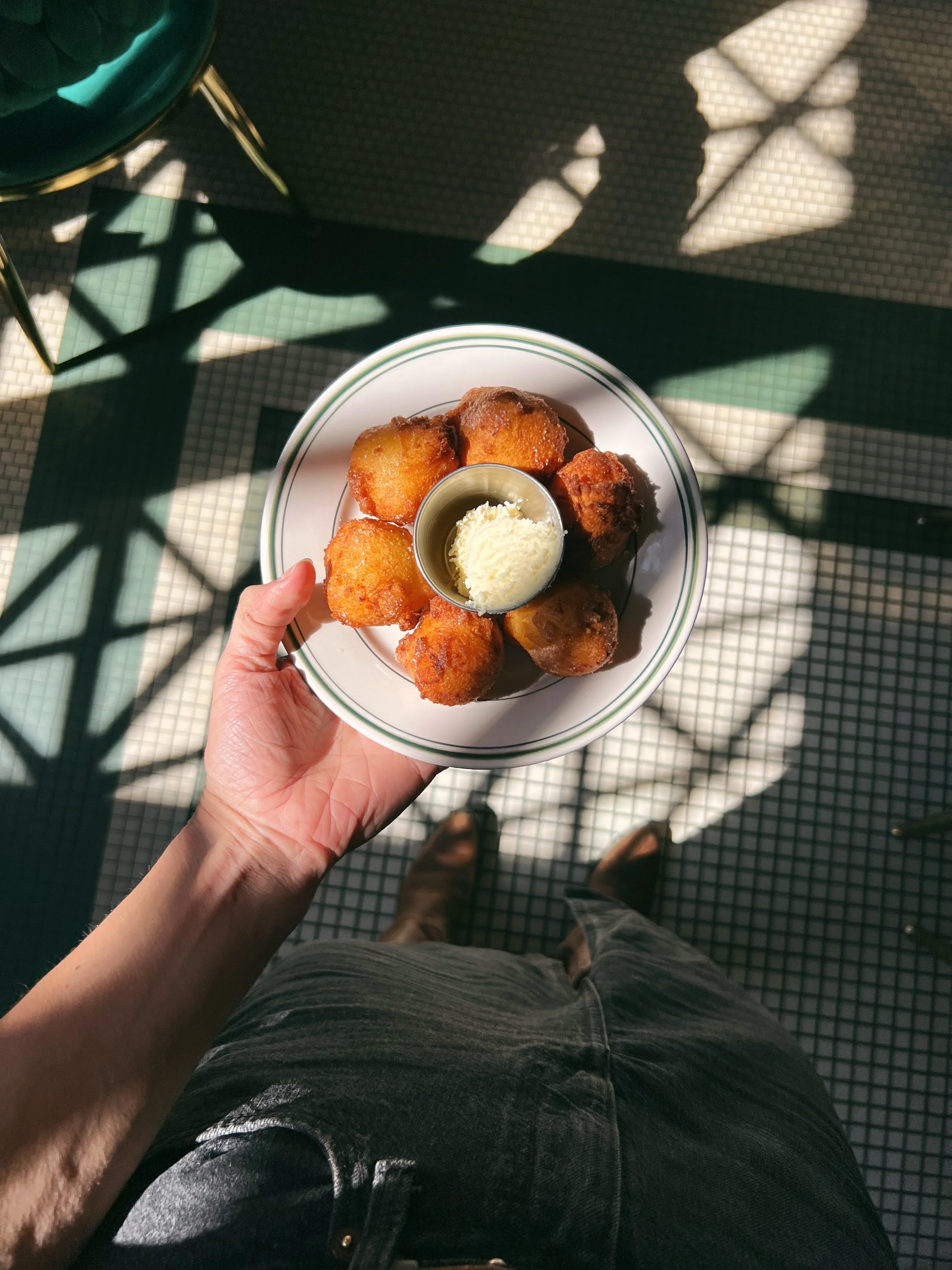 A person holding a plate of fried foods with a cup of butter or margarine in the center, standing on a tiled floor with sunlight and shadows.