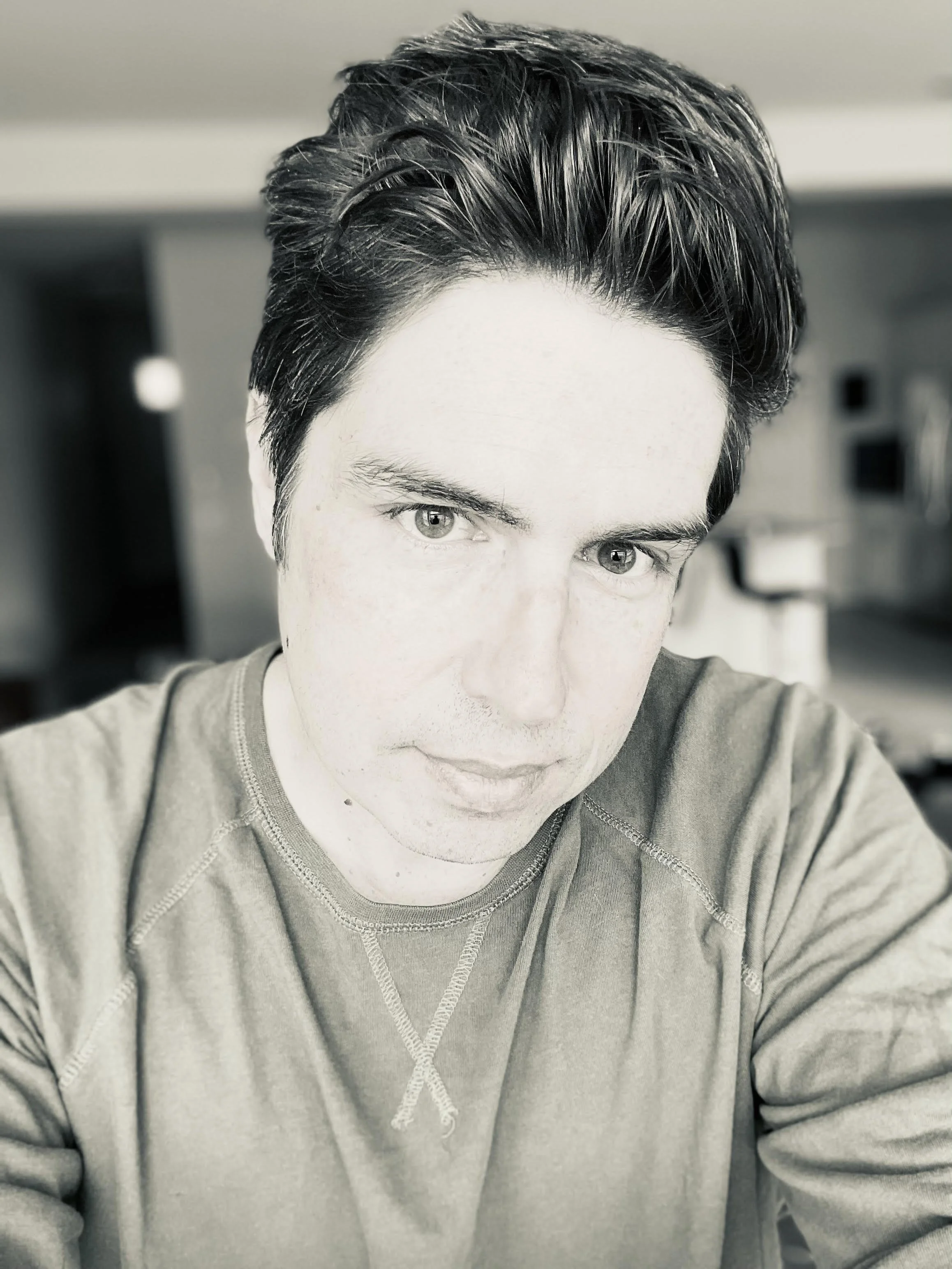 A black and white close-up selfie of a young man with dark, styled hair, light skin, and light-colored eyes, wearing a casual t-shirt, looking directly at the camera with a neutral expression, in an indoor setting.