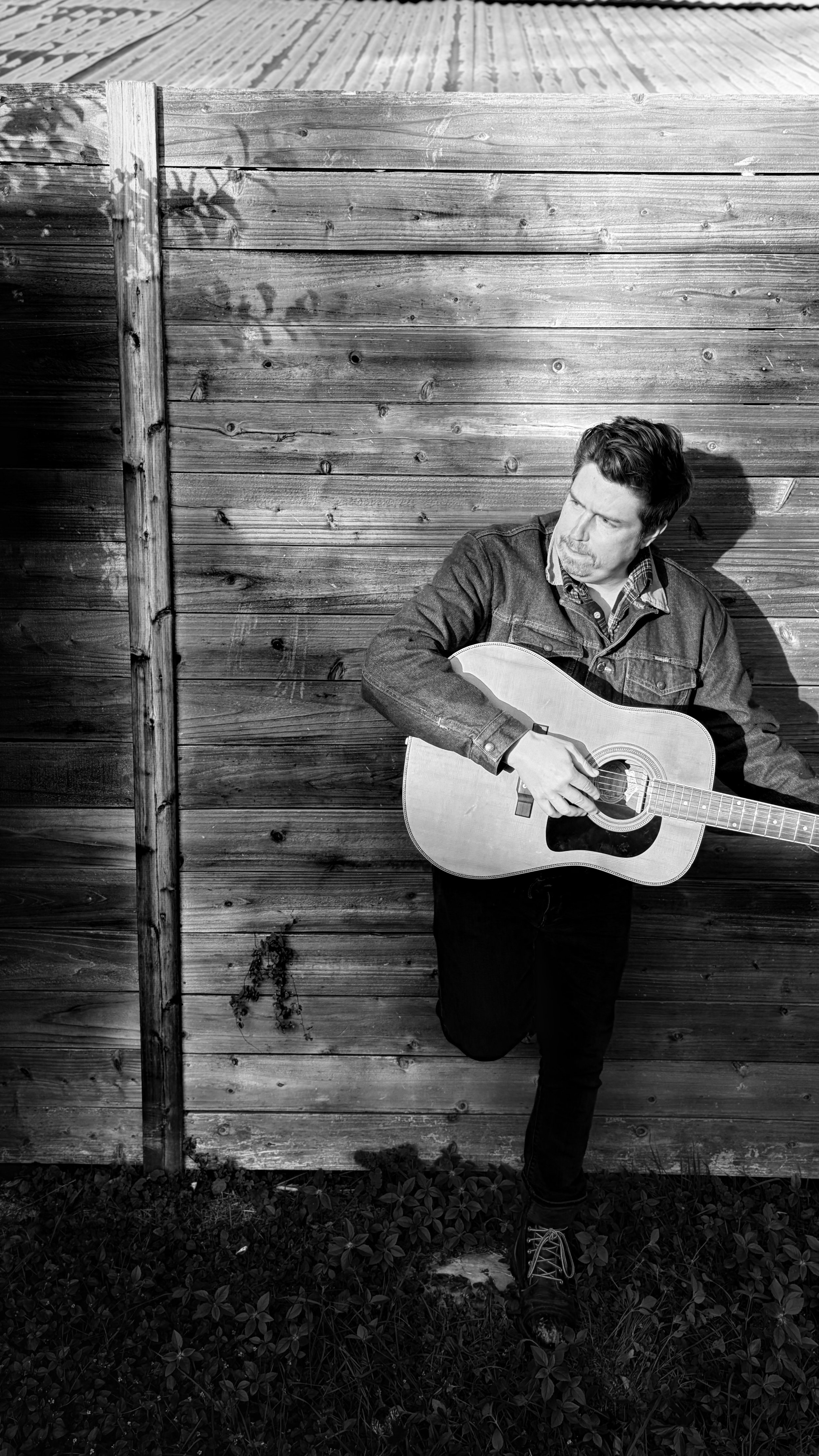 A man playing an acoustic guitar while standing against a wooden fence, with one leg raised and resting on the fence, in black and white.