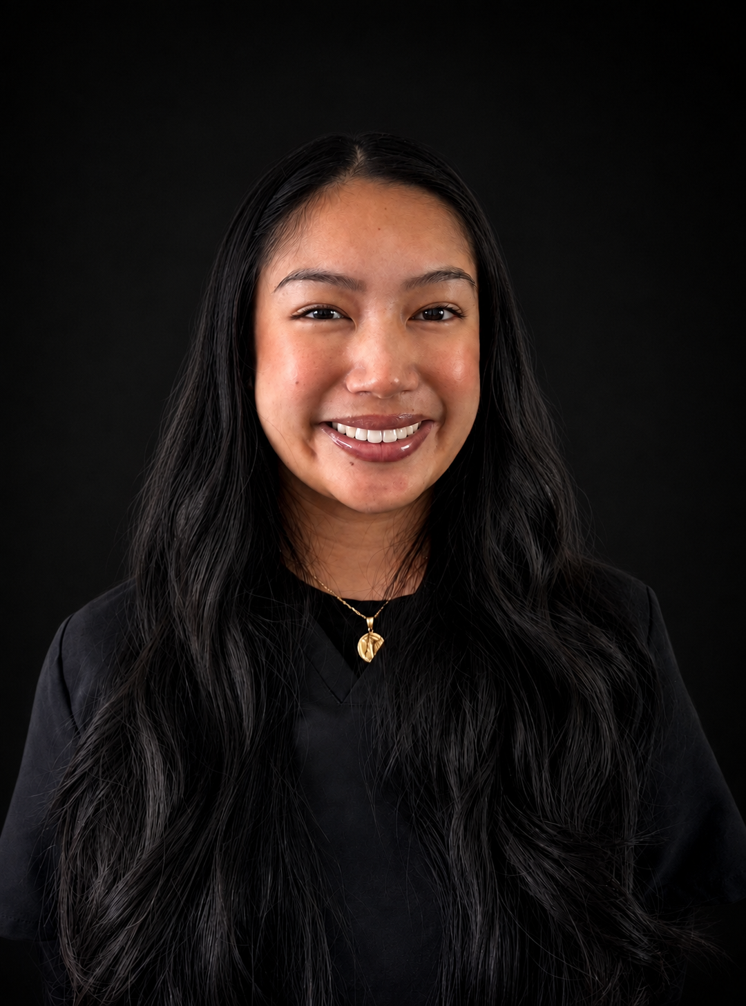 Headshot of a woman with long black hair, smiling, wearing a black top and a gold necklace with a pendant, against a black background.