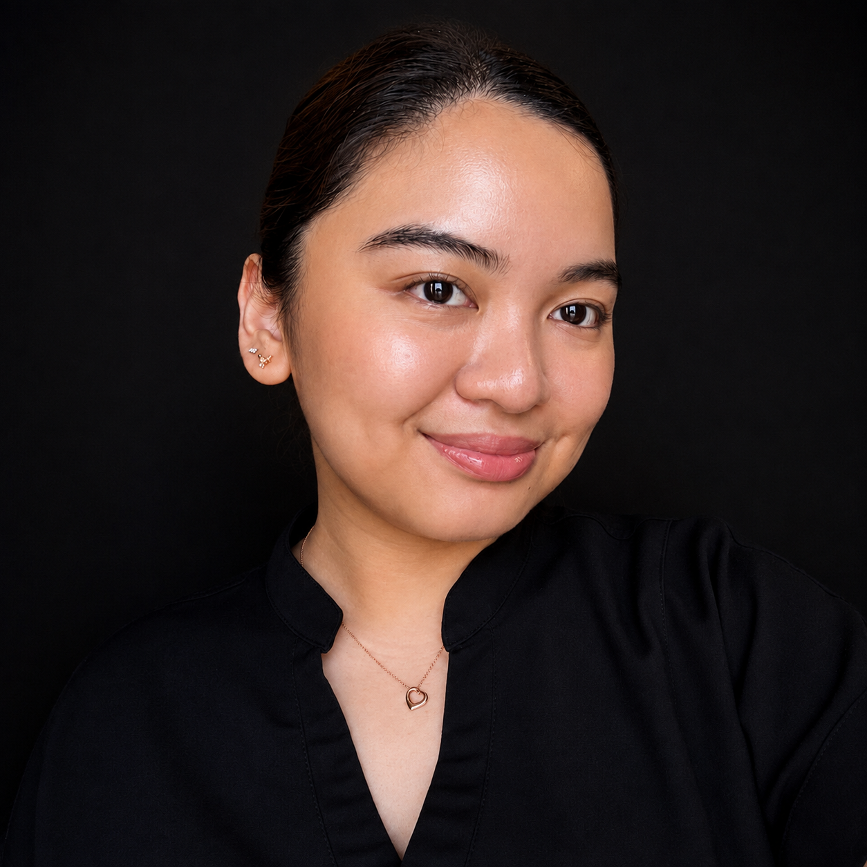 Close-up portrait of a young woman with dark hair, smiling, wearing a black top, a heart-shaped necklace, and small earrings against a black background.