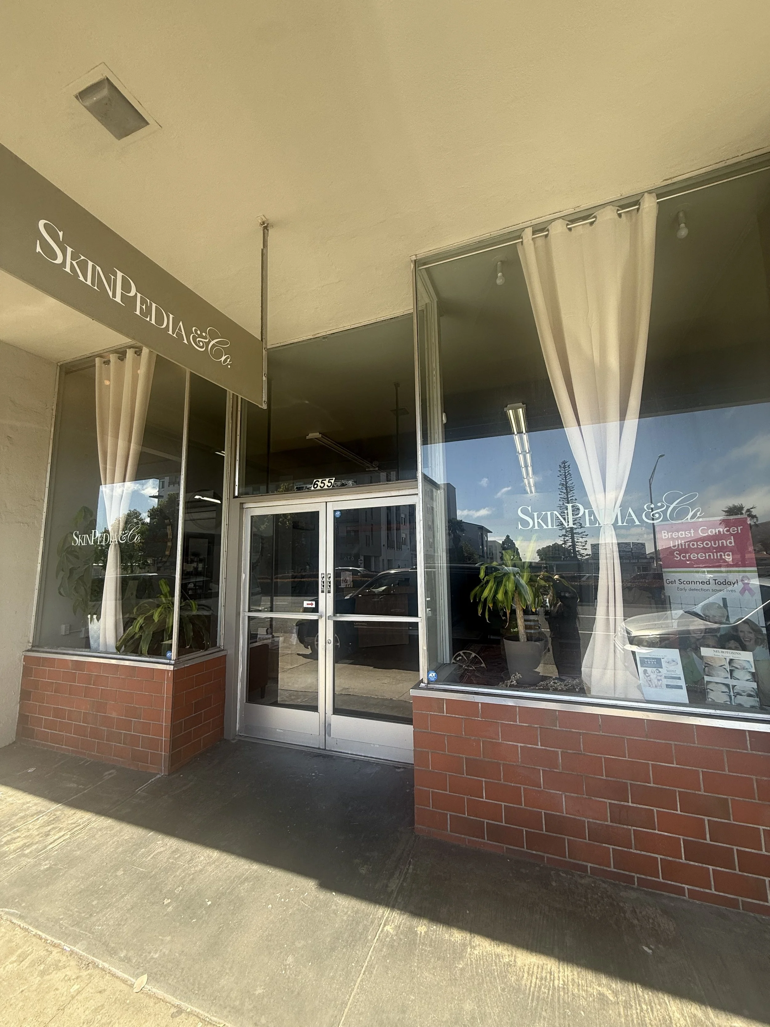 Storefront with large glass windows and door, sign reading 'SkinPedia & Co.', and a pink sign about breast cancer screening inside the window. Two potted plants in front of windows, curtains, and reflections of trees and sky on the glass.