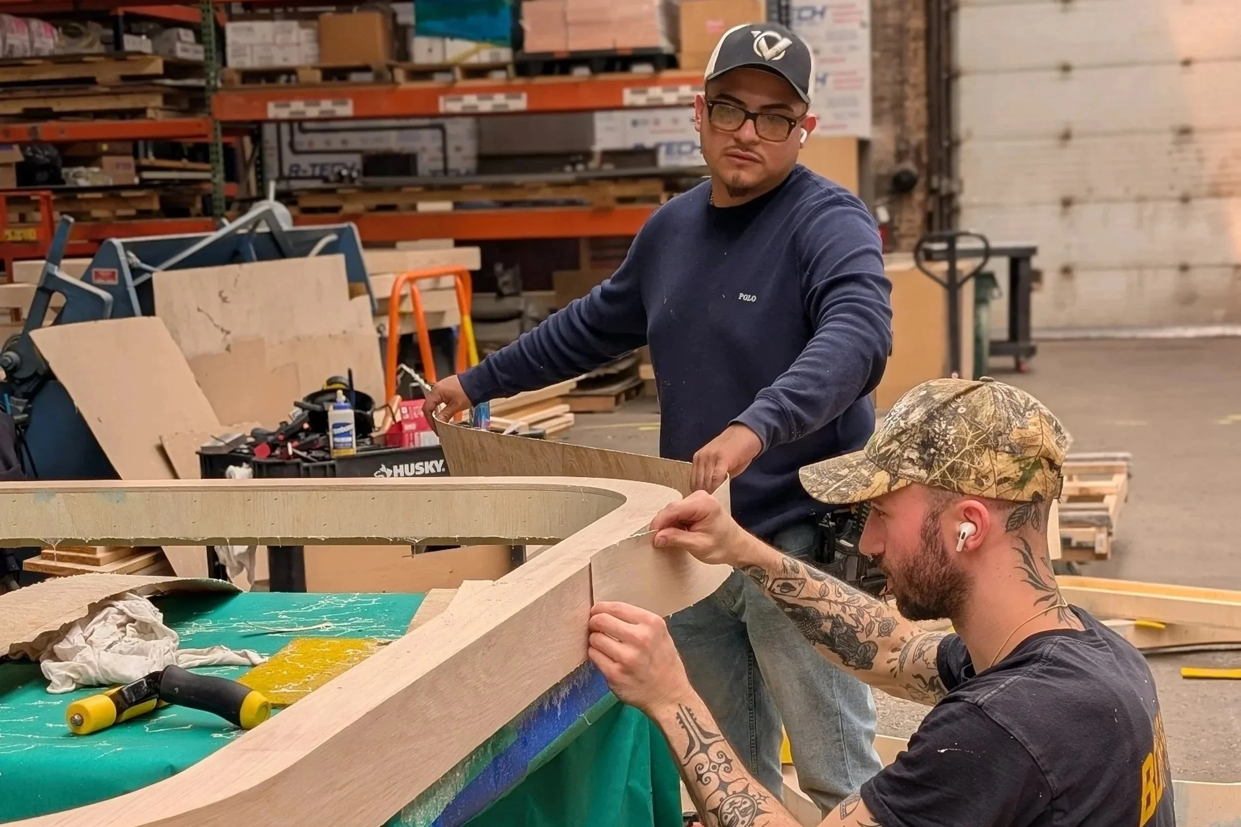 Two men working together on a woodworking project in a warehouse, assembling a large wooden piece, with woodworking tools and materials around them.