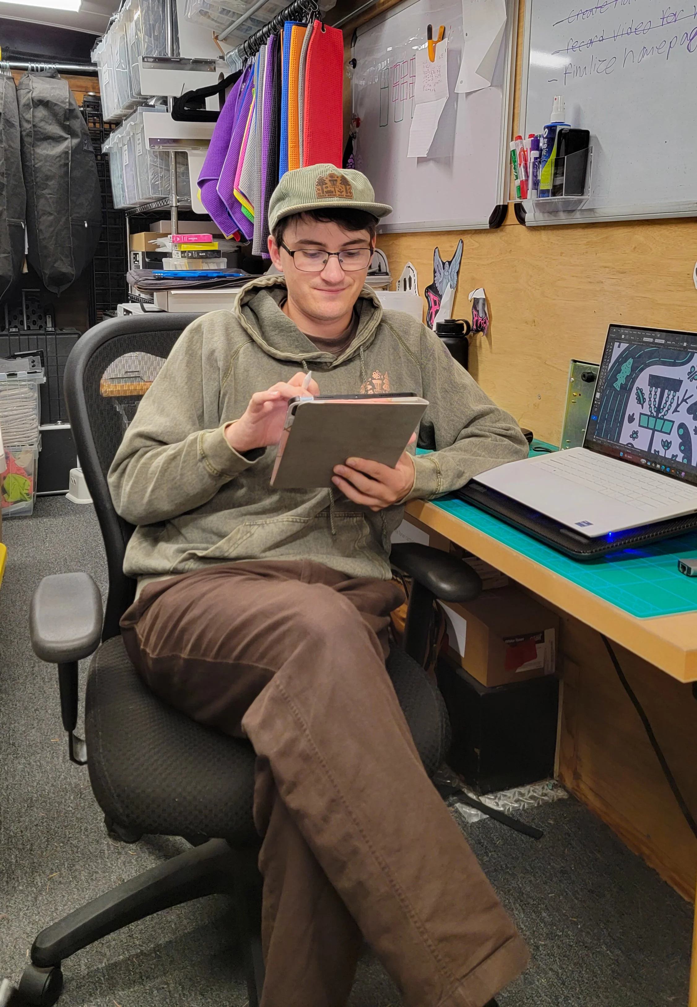 A young man with glasses and a green cap sitting in a black office chair at a desk, looking and smiling at a tablet in his hands. On the desk, there is a laptop, some papers, and a green cutting mat. Behind him are shelves with storage bins and colorful towels hanging. The wall has a whiteboard with notes and drawings and various markers in a holder.