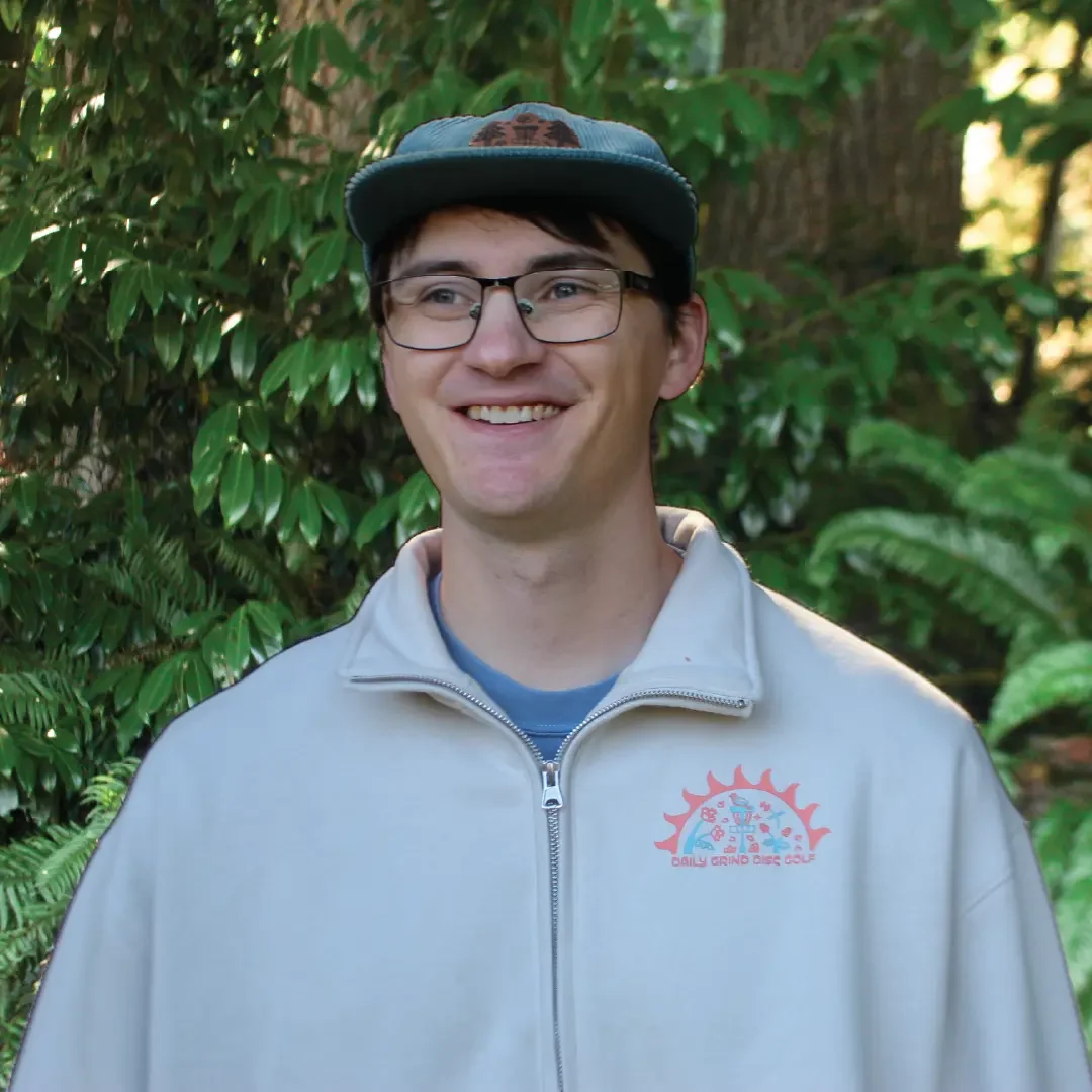 A young man smiling outdoors, wearing glasses, a gray zip-up jacket with a logo on the chest, and a baseball cap, surrounded by green foliage.