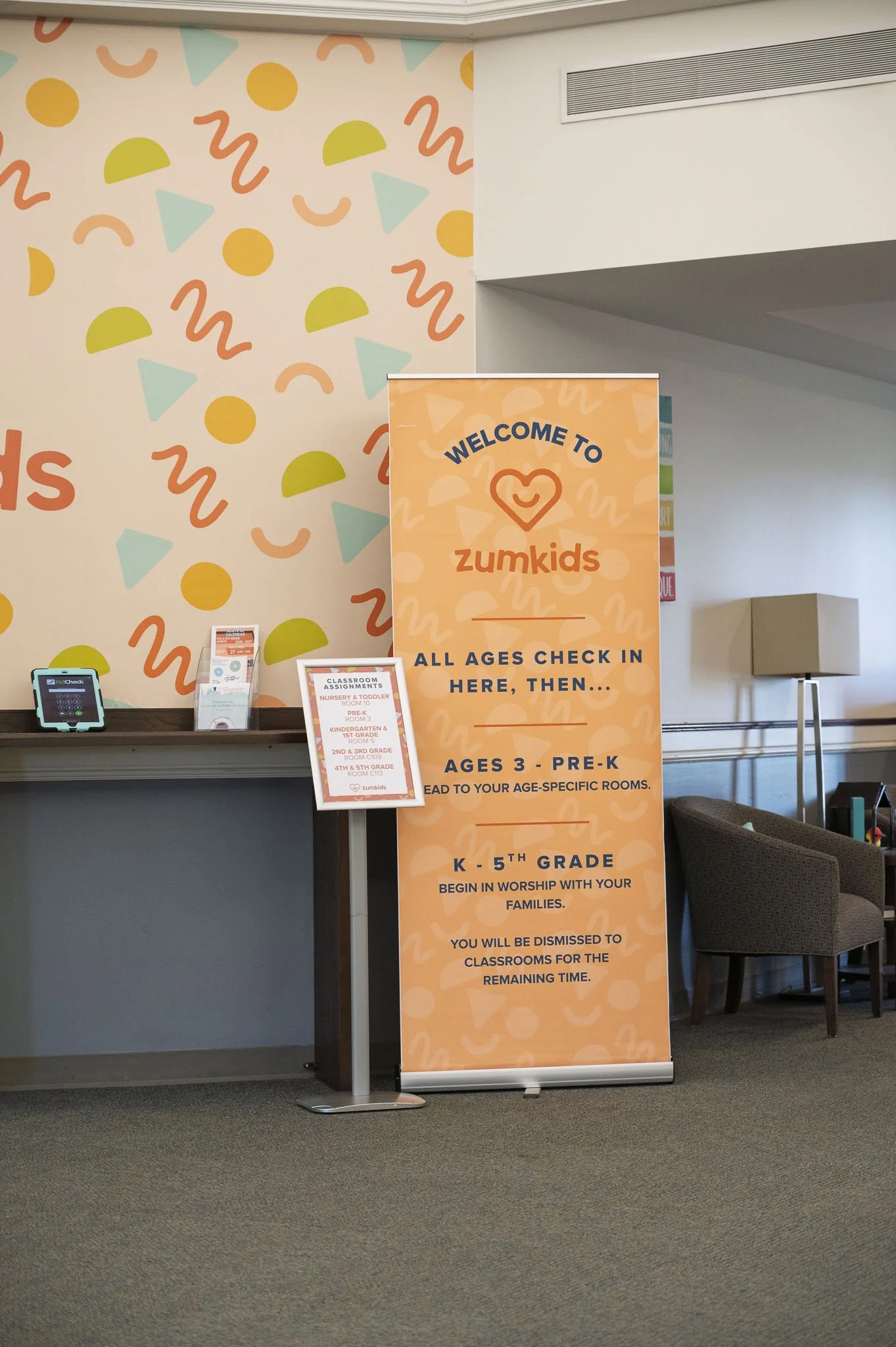 Indoor space with a colorful, geometric-themed wall and a large orange sign for Zumkids, detailing check-in procedures for children ages 3 through 5th grade, with chairs and a small table nearby.