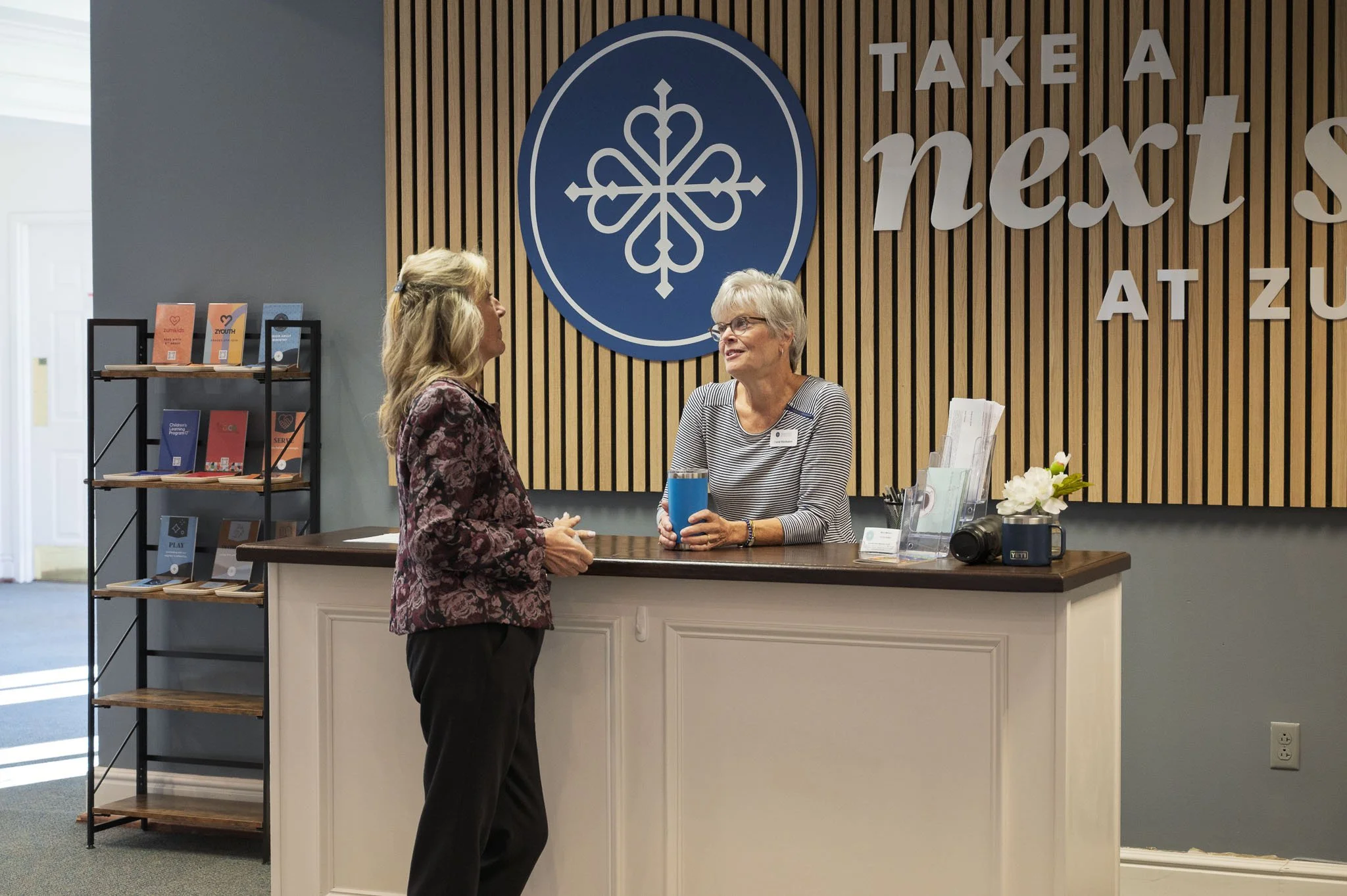 Two women engage in conversation at a reception desk within a wellness or health center, featuring a modern wooden slat wall with a large blue emblem and the words 'Take a Next Step At ZU' surrounding it.