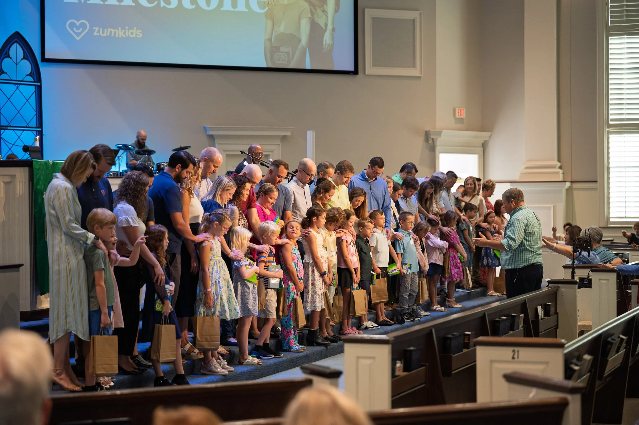 Group of children and adults standing on stage during a church service, holding hands and praying. A man is leading the prayer, and there are musical instruments and a large screen with a presentation in the background.
