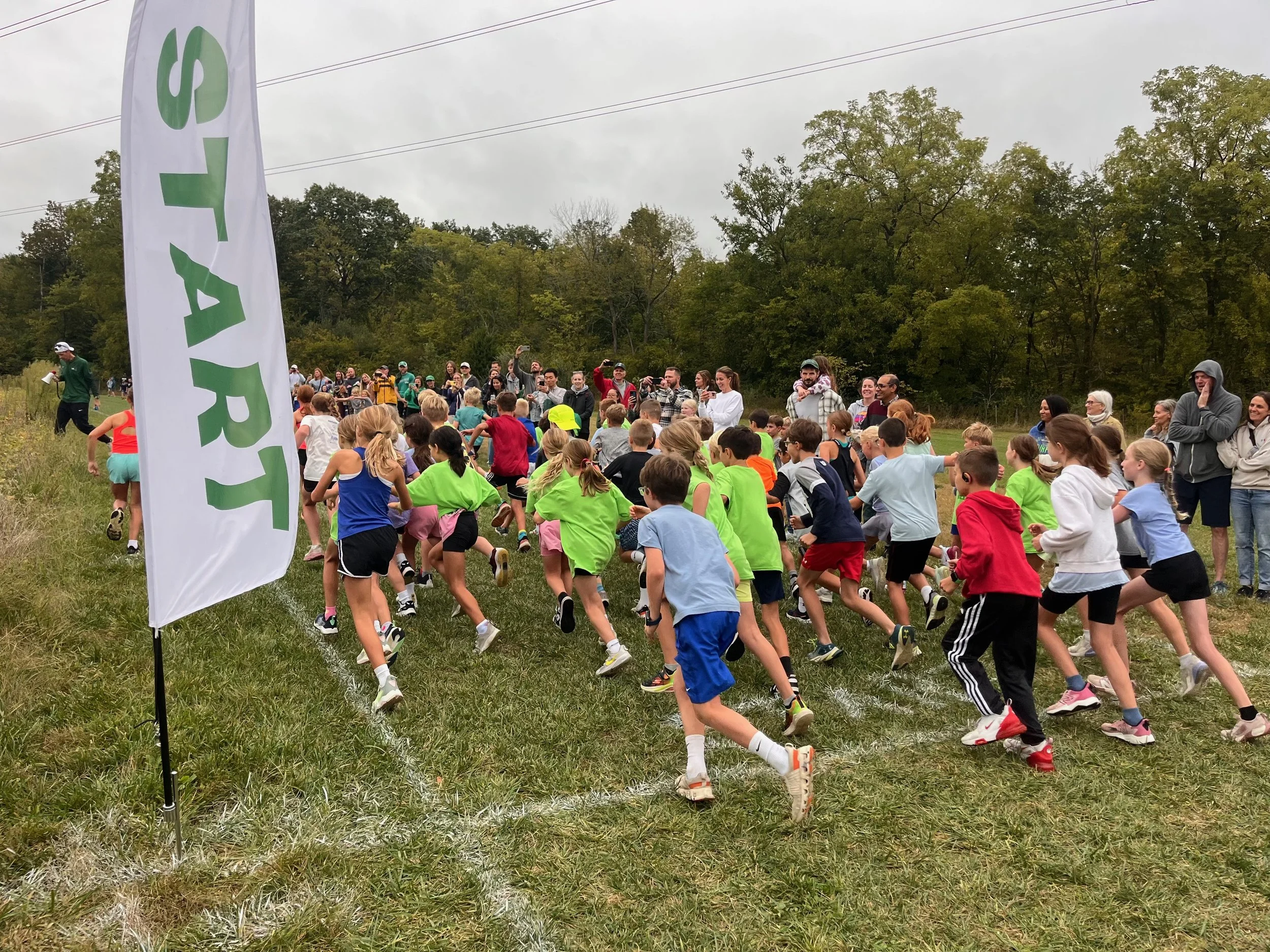 Children and adults participating in an outdoor running event on a grassy field, with many people taking photos and a large flag with green text in the foreground. Trees and overcast sky are in the background.