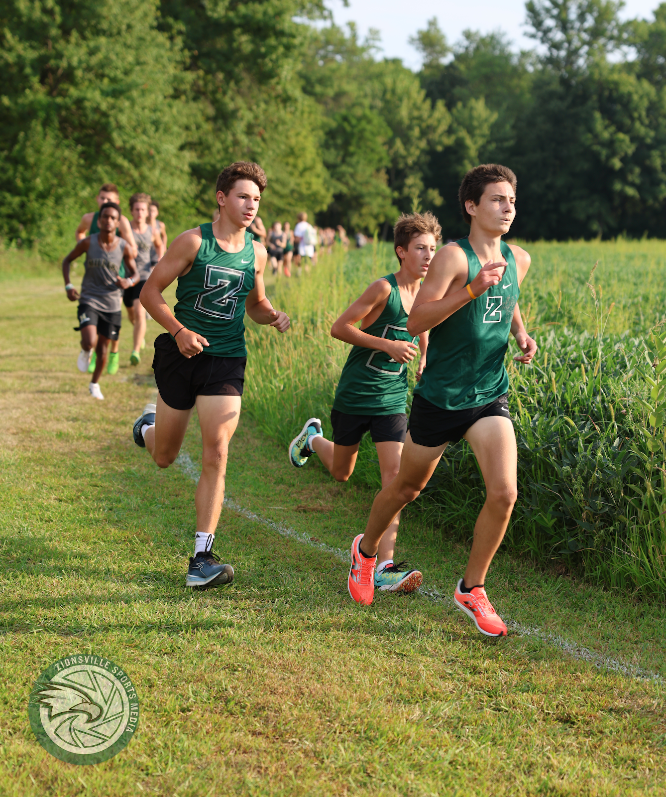 Group of young male cross-country runners wearing green uniforms running along a grassy trail beside a field with green trees in the background.