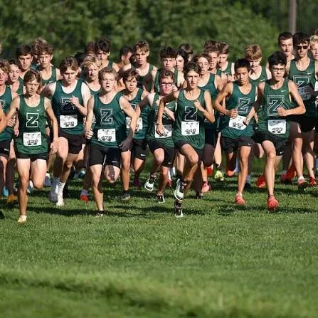 Group of male cross-country runners wearing green and black uniforms at the start of a race on a grassy field.