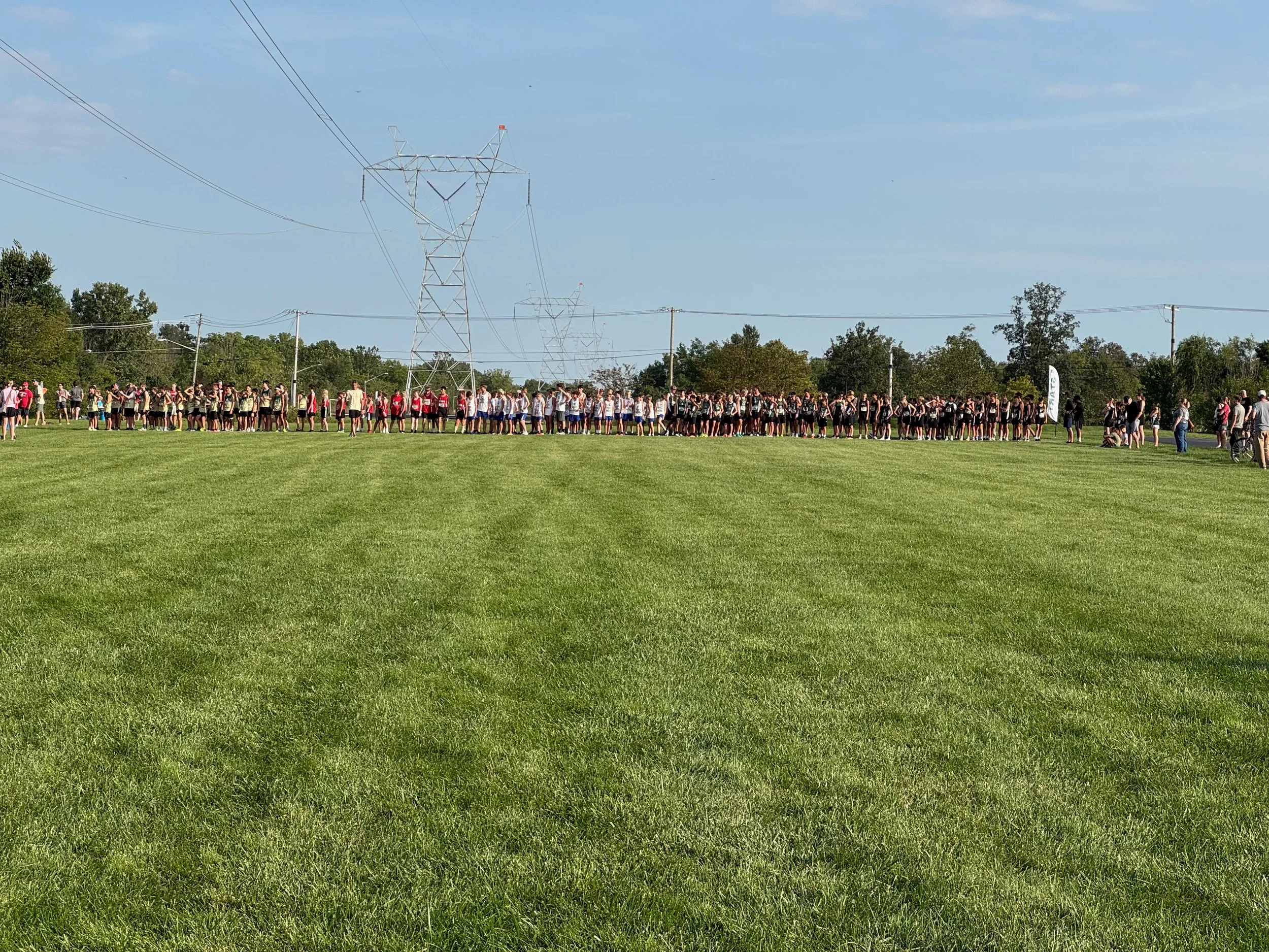 A large group of people, possibly a sports team, standing in multiple rows on a grassy field. Power lines and utility poles are visible overhead, with trees and a blue sky in the background.