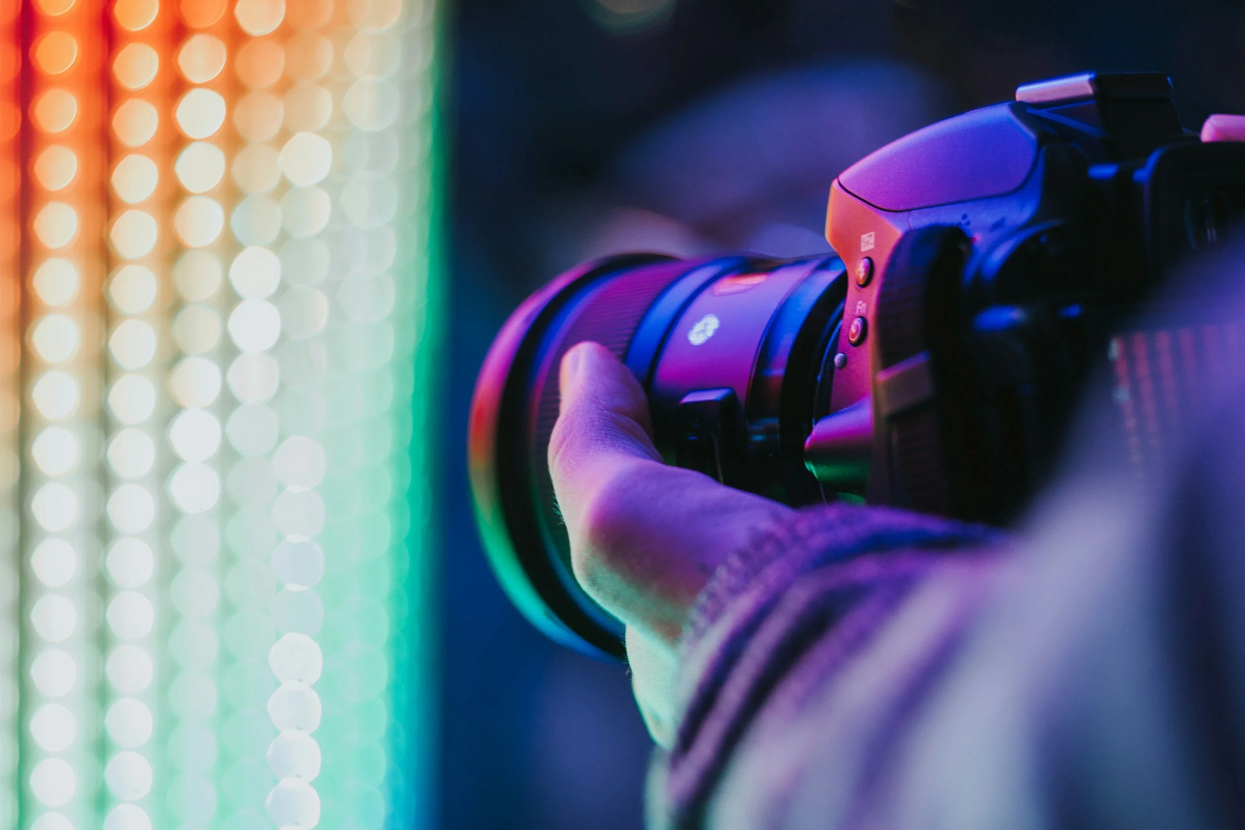 Close-up of a digital camera being held by a person's right hand, aimed at a colorful LED light background with bokeh effect.