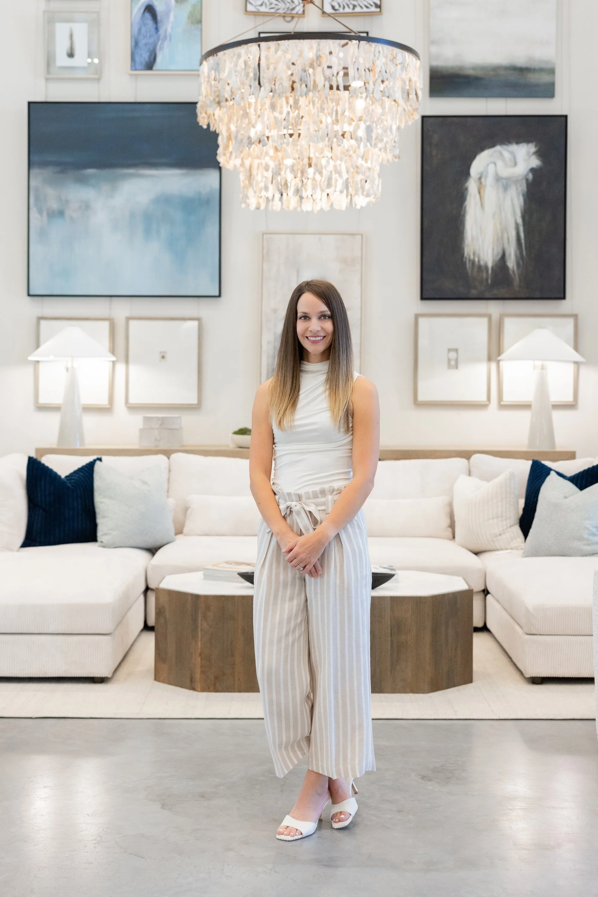 A woman with long brown hair smiling, standing in a modern living room with a white couch and framed artwork on the wall behind her, a large chandelier hanging from the ceiling, and white table lamps on either side of her.