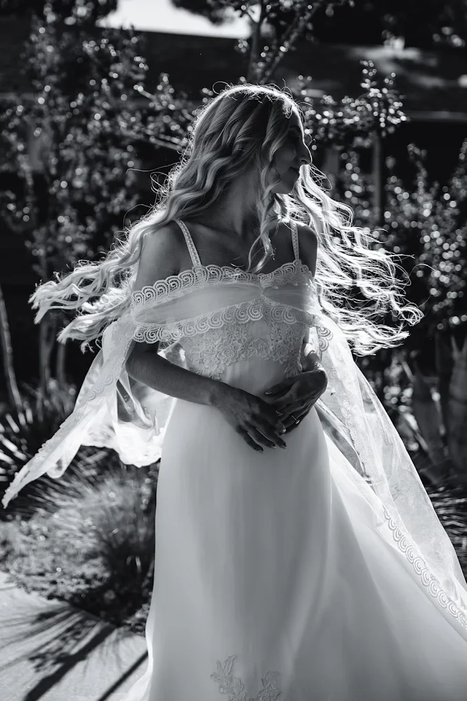 Black and white photo of a woman in a flowing wedding dress with lace details, standing outdoors with sunlight illuminating her hair and dress, surrounded by trees and plants.