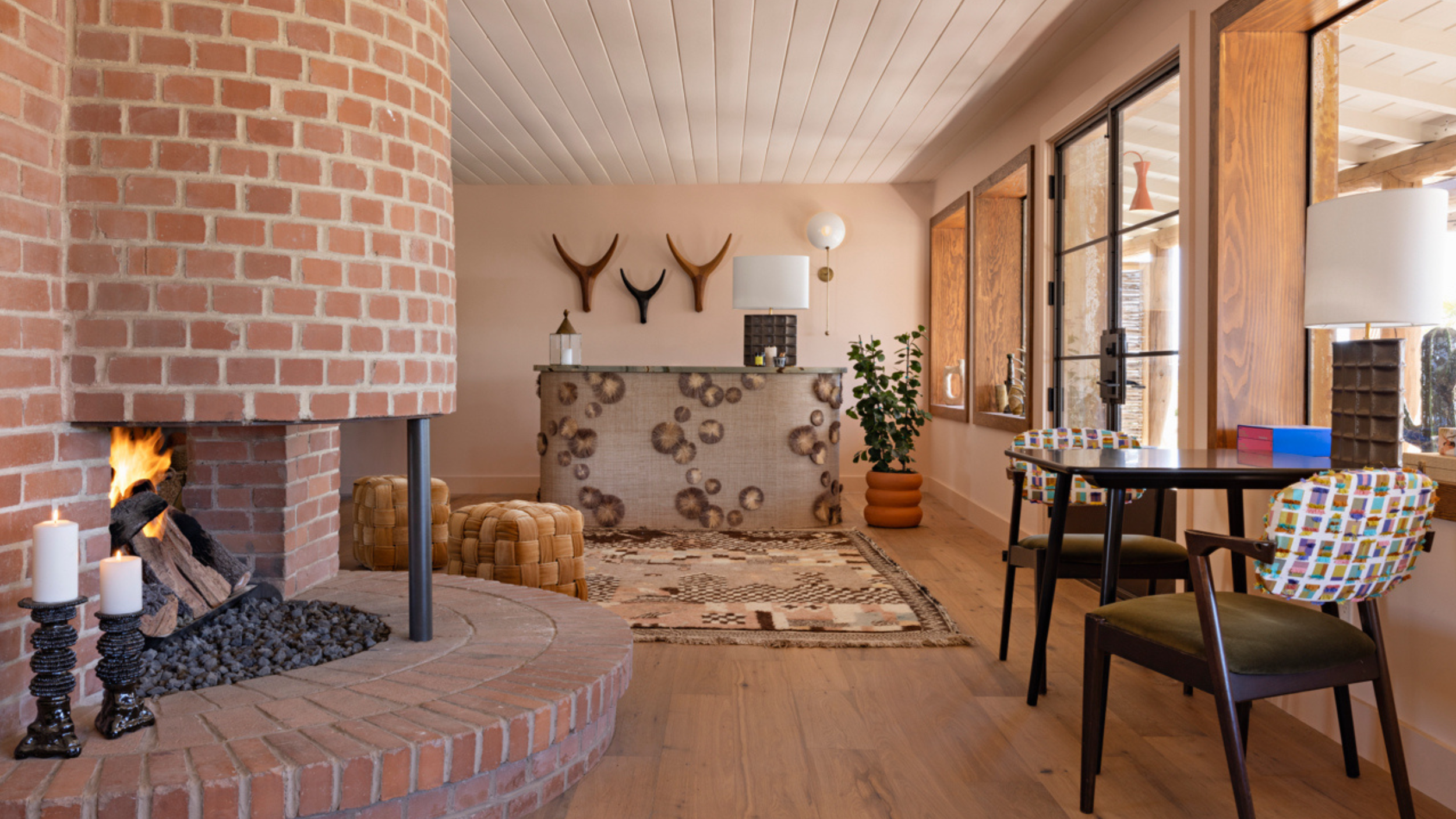 A cozy living room with a brick fireplace, decorative antler wall art, a patterned area rug, and a wooden table with chairs featuring colorful patterned upholstery. There are large windows and a potted plant near the sofa.