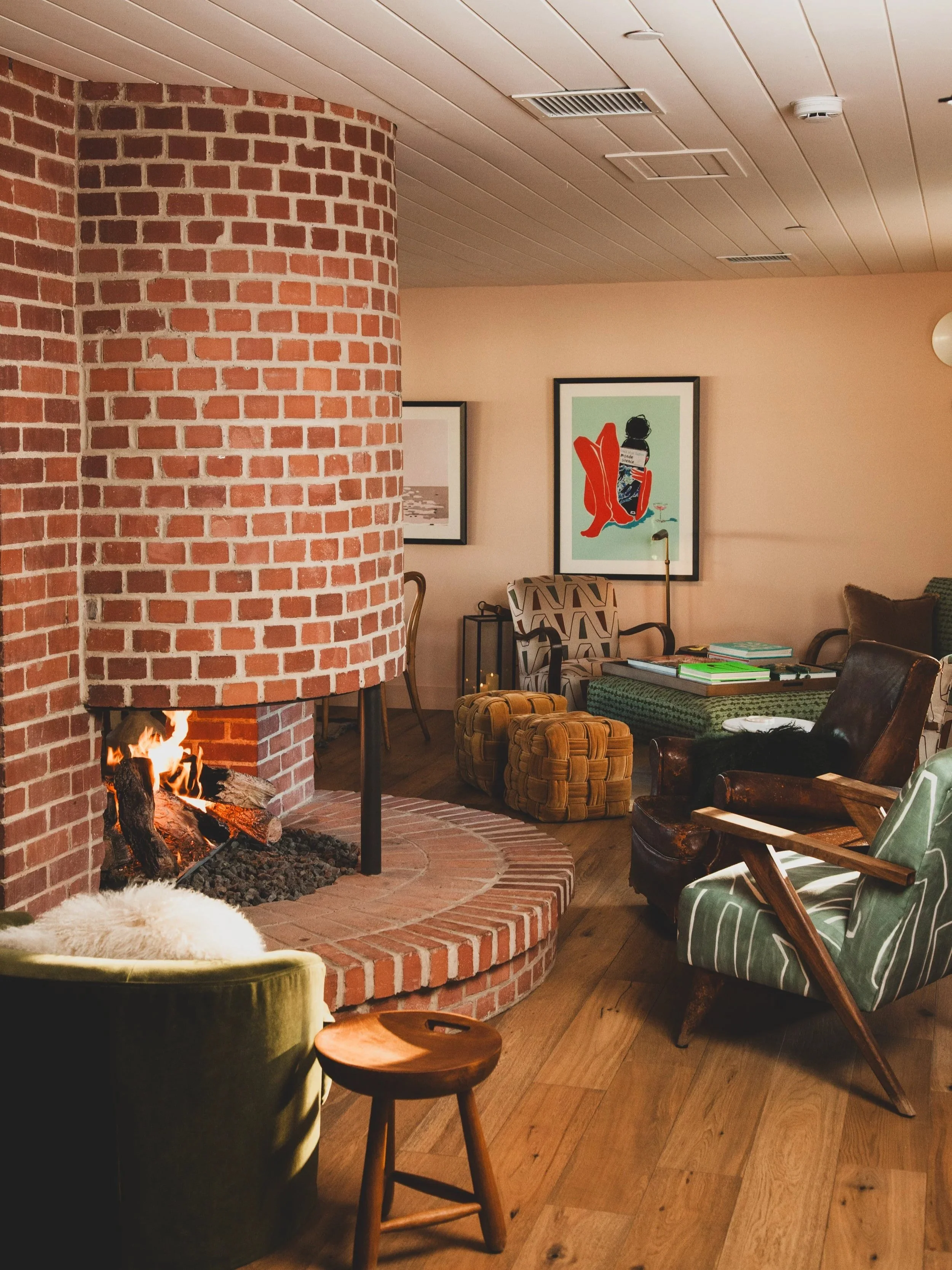 A cozy living room with a brick fireplace, a fire burning, surrounded by various chairs and tables, and framed art on the walls.