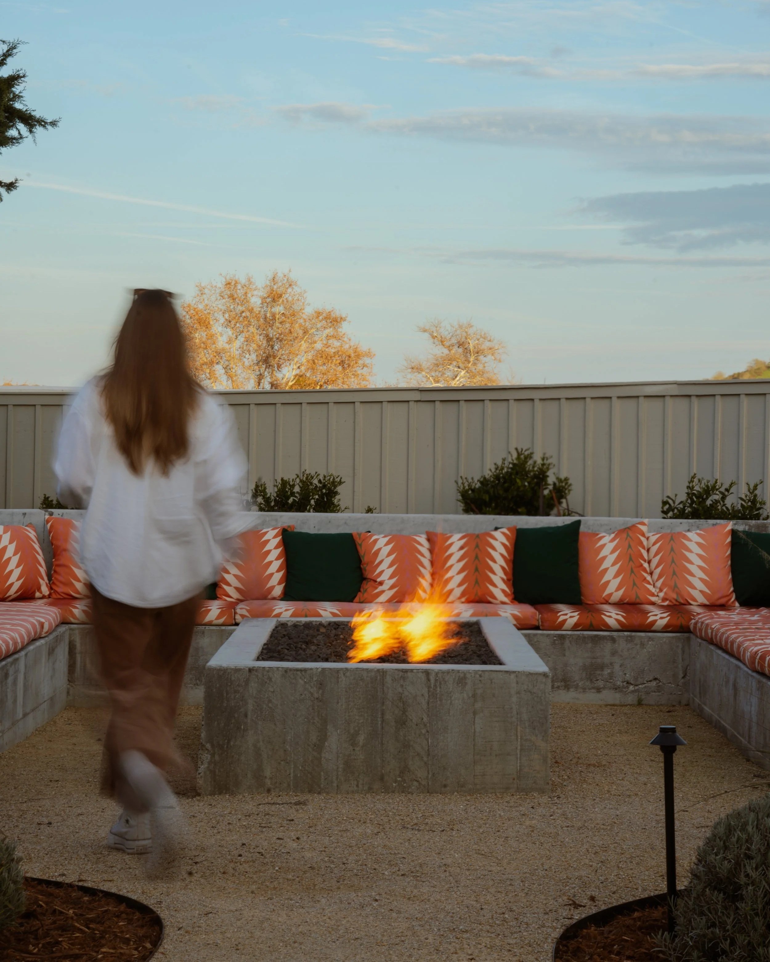 A person with long reddish hair wearing a white jacket and beige pants walking past a rectangular outdoor fire pit with flames. In the background, there are orange and green cushioned seating along a curved concrete wall, and a backyard with a white 