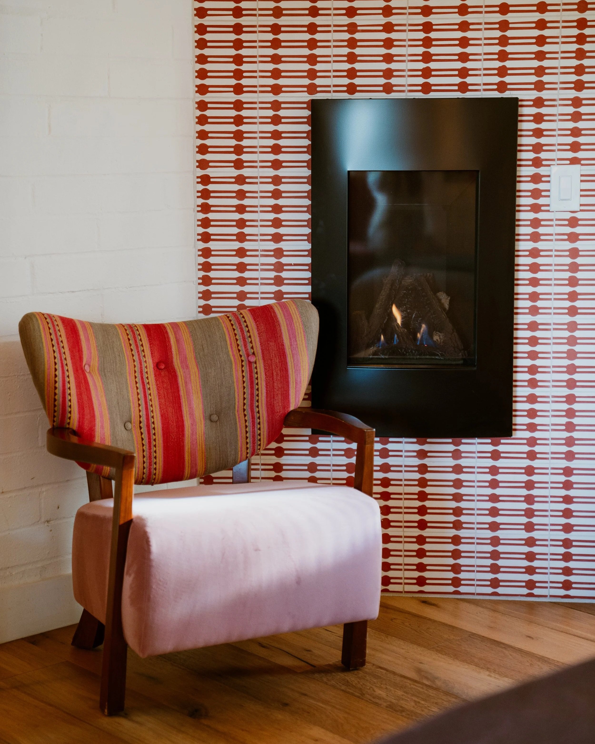 A living room corner with a pink upholstered armchair, a small wooden side table, and a fireplace with a black frame. The wall behind features a red and white patterned tile design on the right and a plain white brick wall on the left. A white wall s