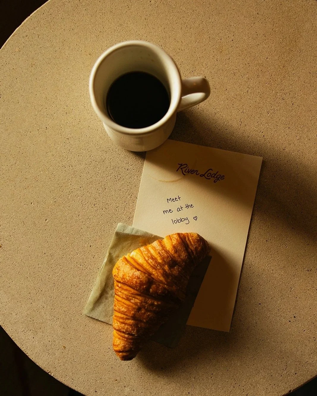 A cup of black coffee, a croissant on a napkin, and a handwritten note on a table. The note says 'River Lodge, Meet me at the lobby' with a heart symbol.