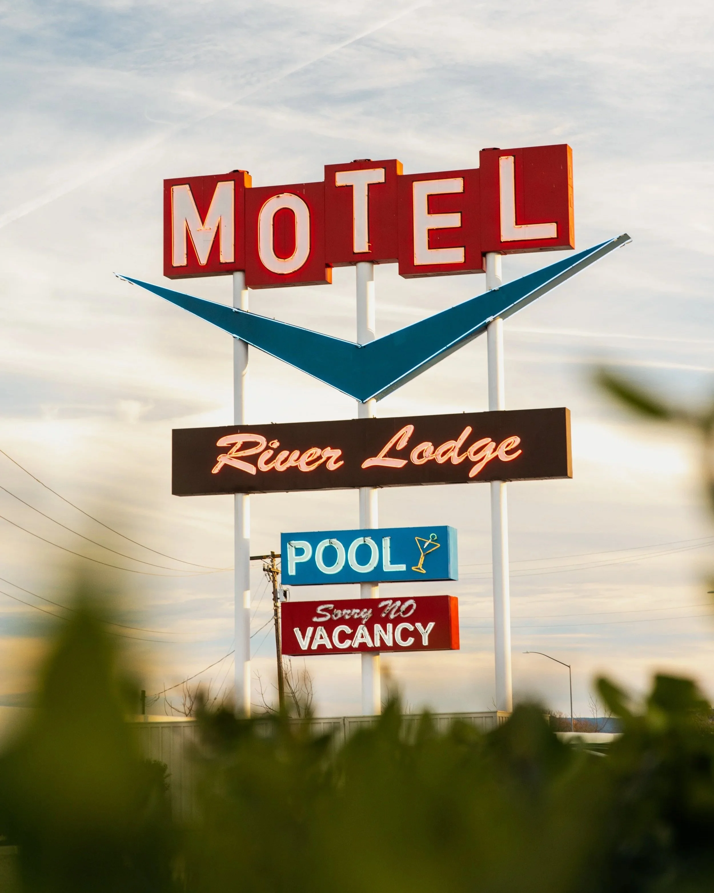 Sign with the words 'Motel' in large neon letters, featuring a blue arrow and smaller signs for 'River Lodge,' a pool, and a no vacancy notice.