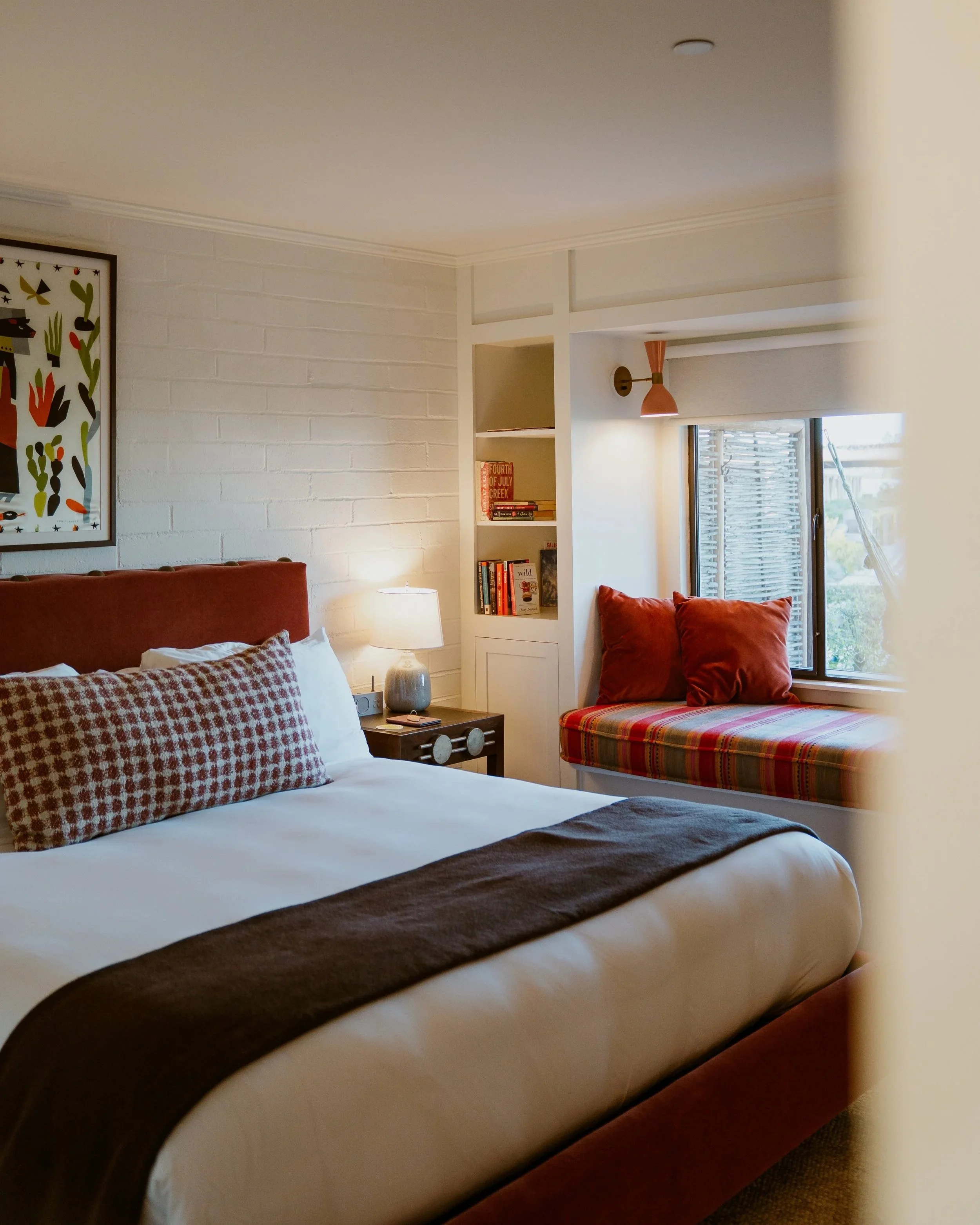 A cozy bedroom with a bed, a nightstand, a table lamp, a built-in bookshelf, and a window with a view outside. The room has white walls and warm-toned cushions.