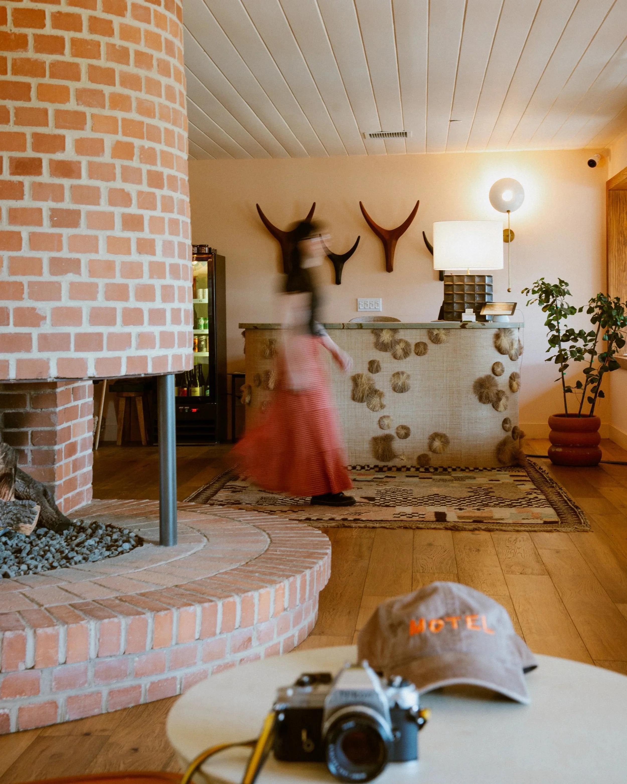 A room with a brick fireplace on the left, wooden floor, and a low table in the foreground with a camera on it, labeled 'MOTEL' on its cover. There are wall-mounted deer antlers and a lamp on a side table, plants, and an open cupboard in the backgrou