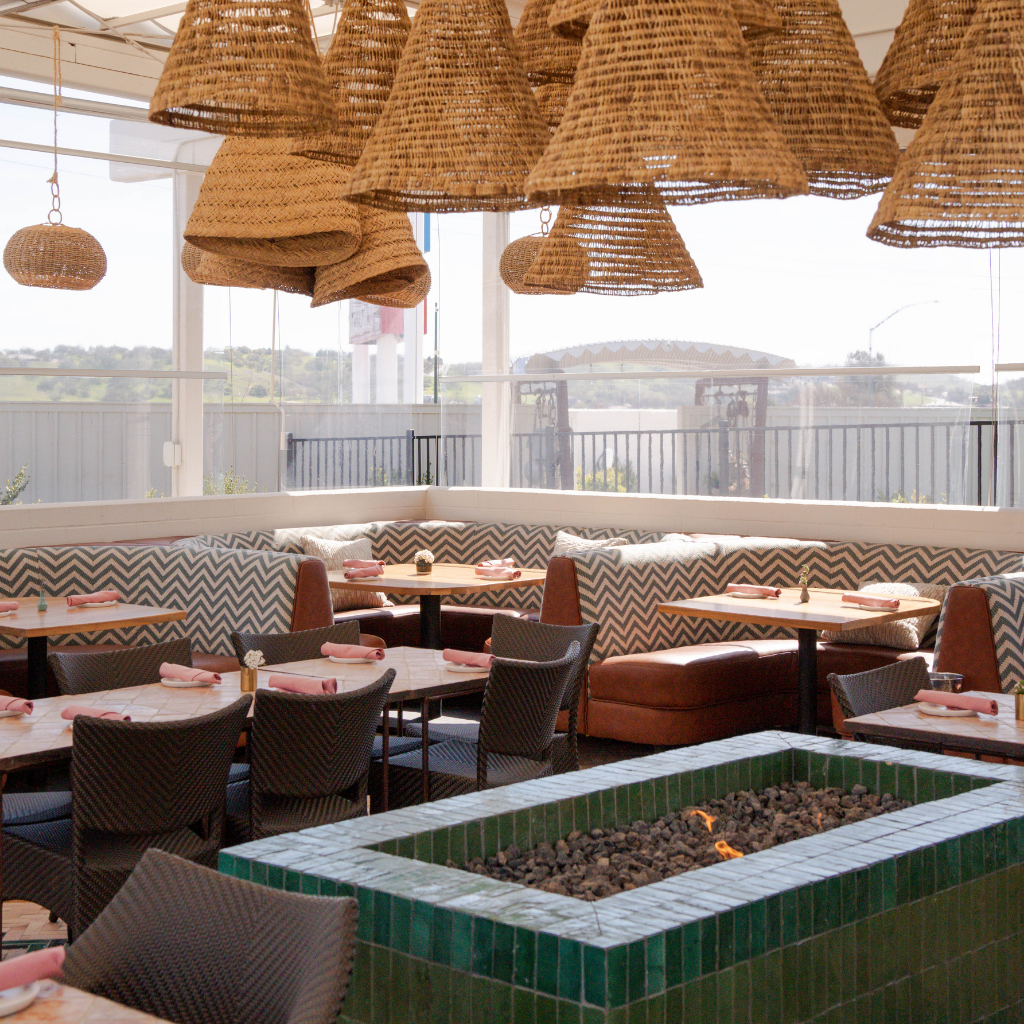 Interior of a restaurant with wicker pendant lights hanging from the ceiling, tables set with pink napkins, and a built-in seating area with chevron-patterned cushions. Large windows reveal an outdoor view with a railing and distant hills, and a rectangular fire pit with rocks is in the foreground.
