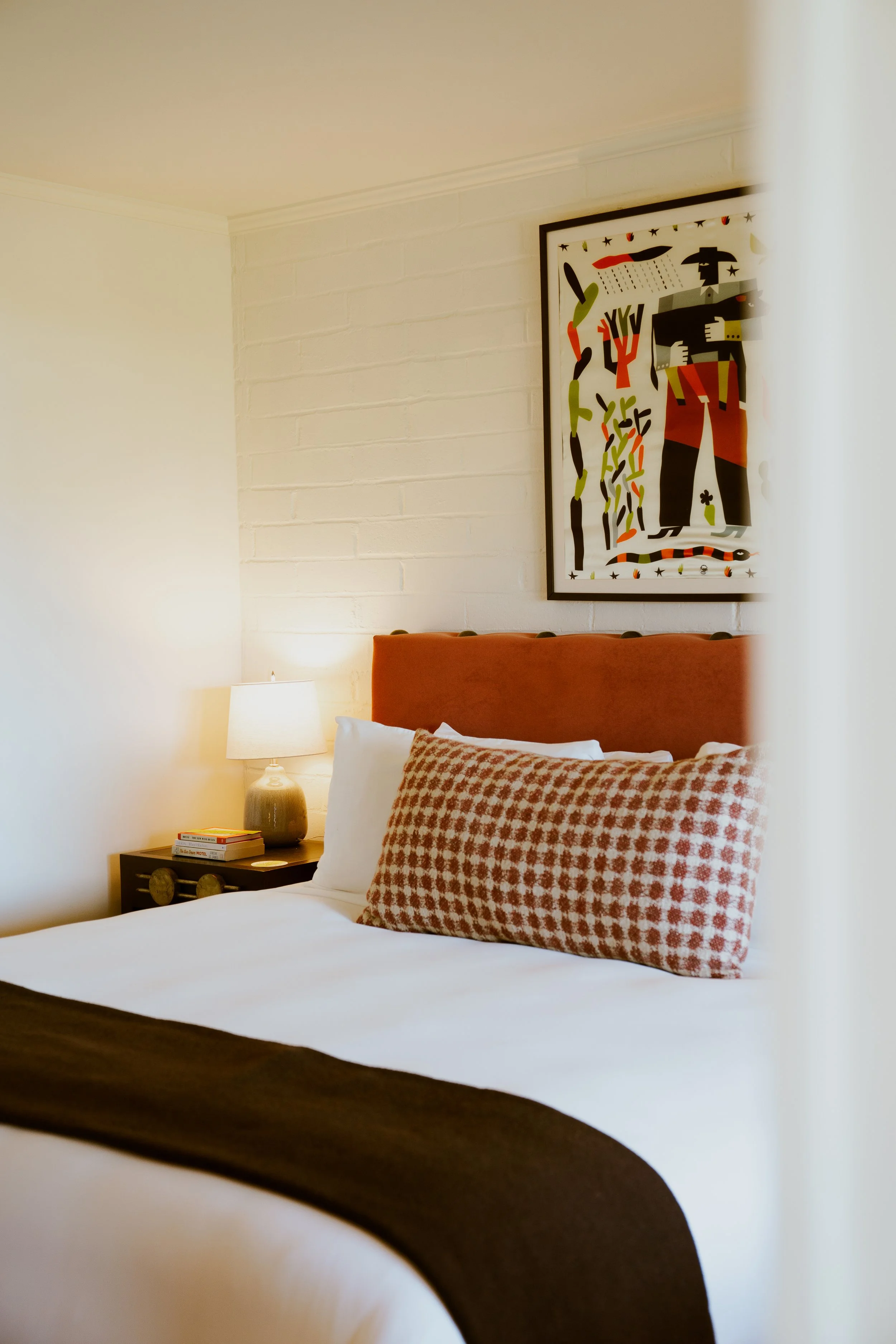 A bedroom with a bed featuring red patterned pillows, a nightstand with books, a table lamp, and framed abstract artwork on a brick wall.