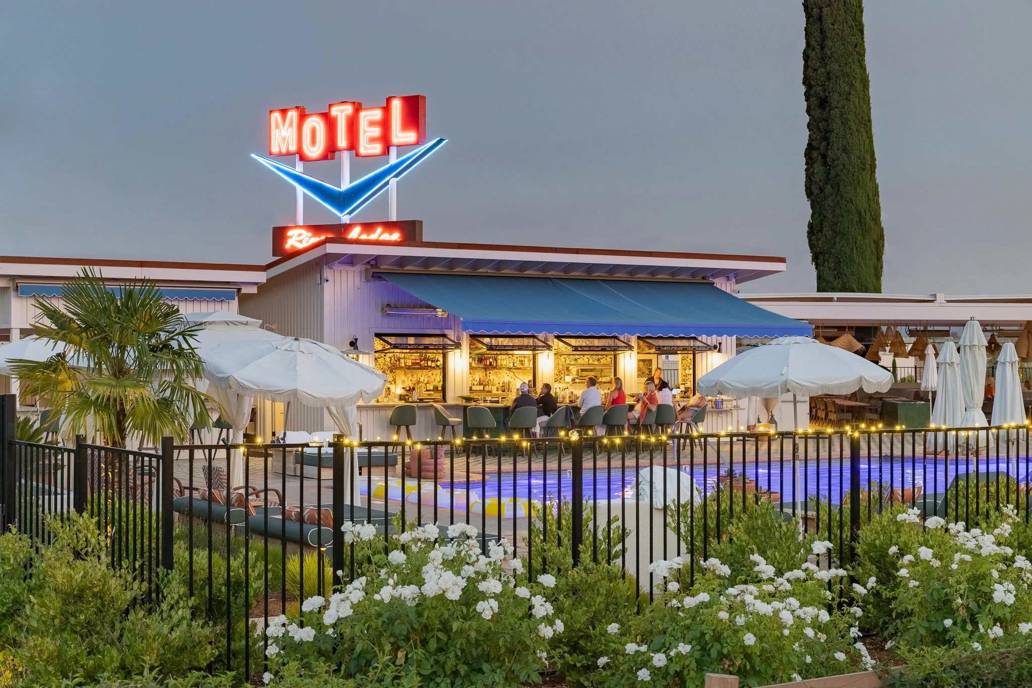 Nighttime scene of a motel with neon sign, outdoor seating area, swimming pool, and surrounding greenery.
