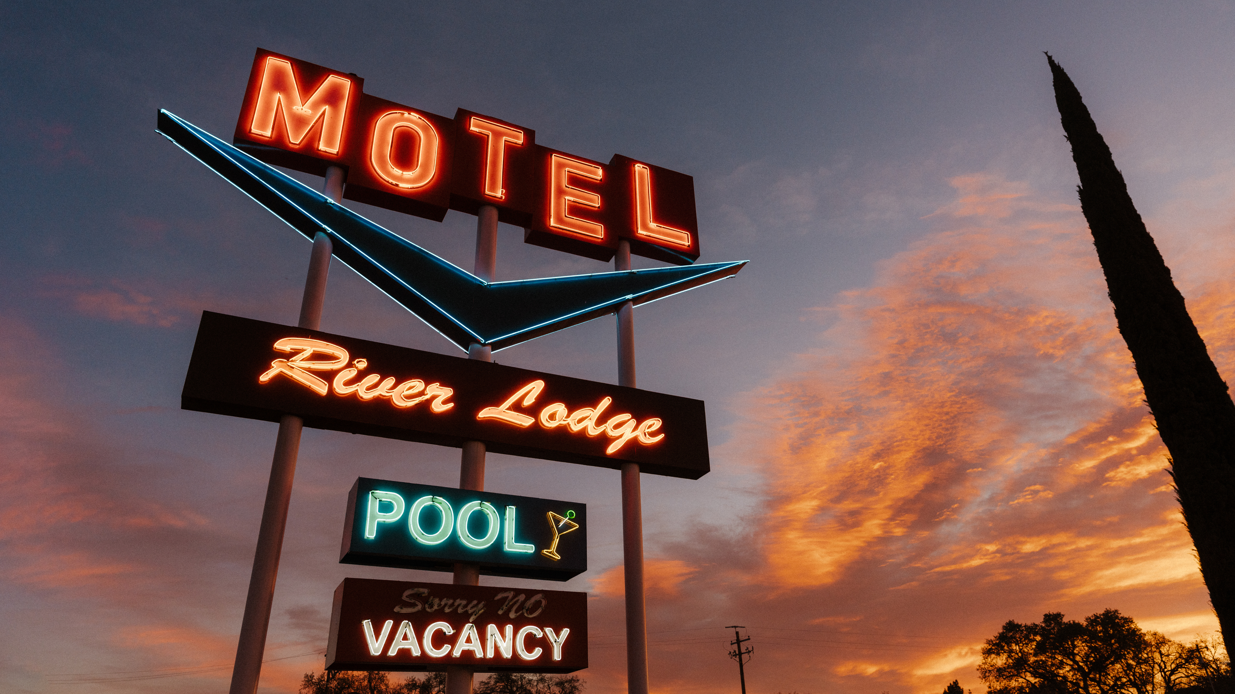 A neon sign for a motel and river lodge against a sunset sky, with additional signs for a pool, sorry no vacancy, and a martini glass.