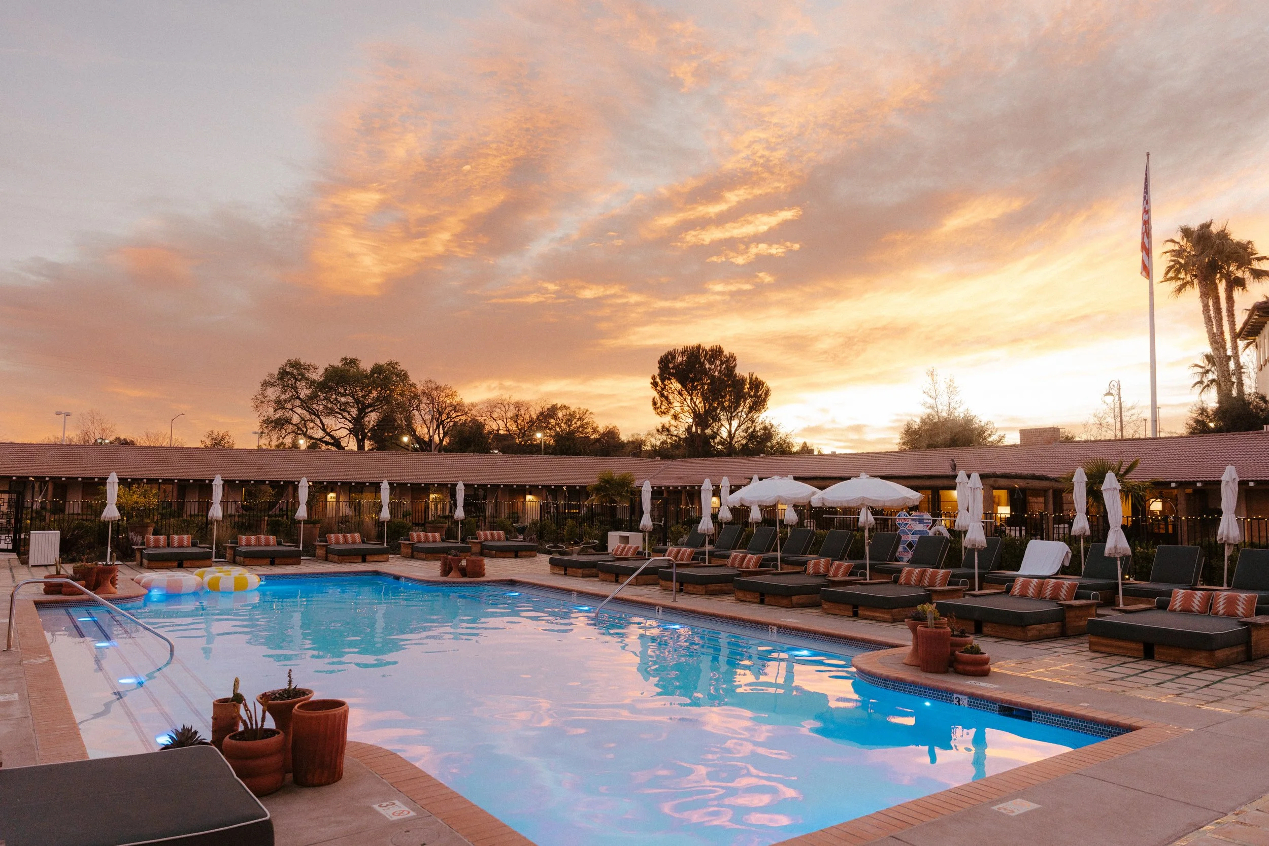 A swimming pool at sunset with lounge chairs, umbrellas, and potted plants around it, with trees, a building, and an American flag in the background.