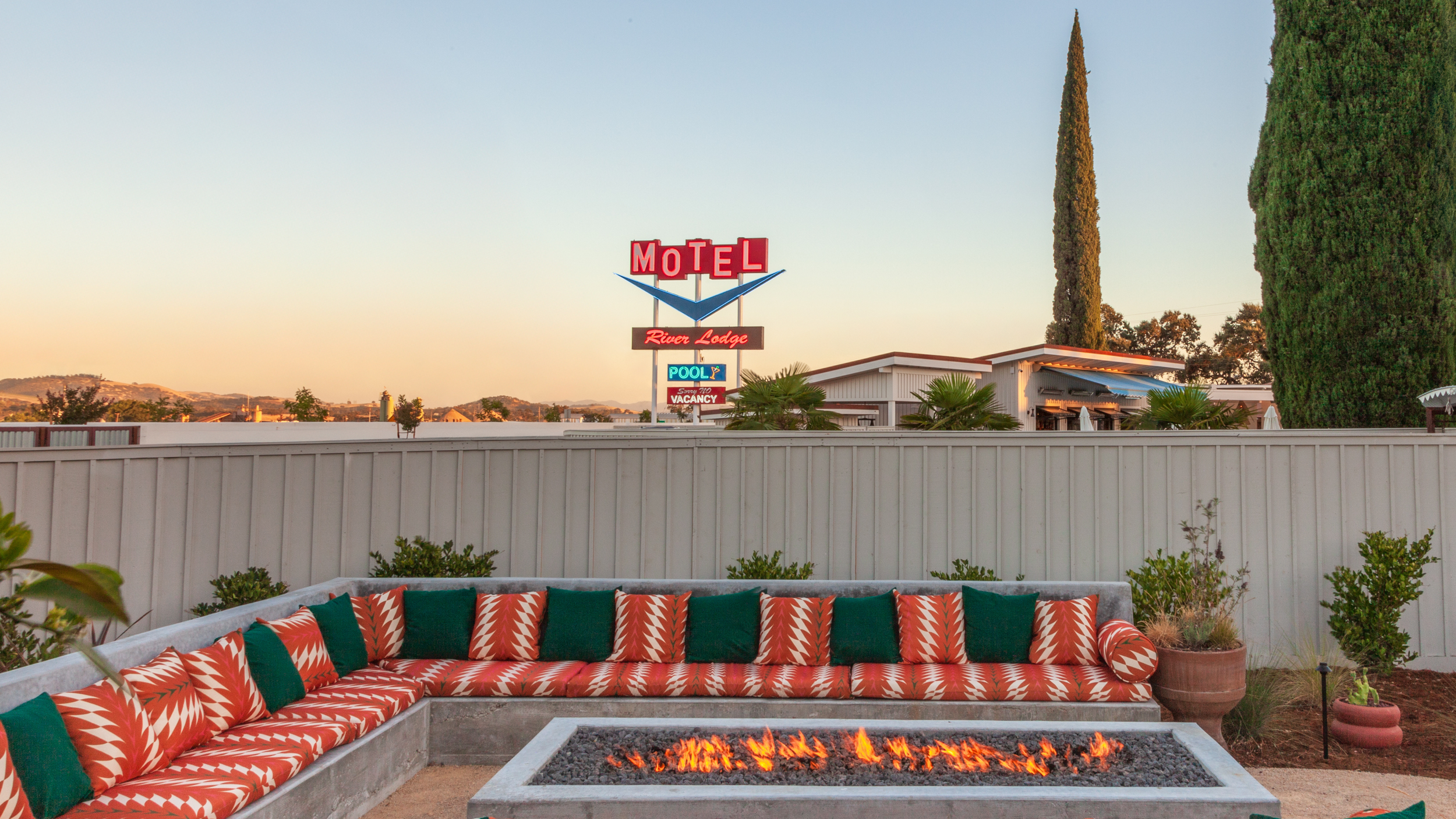 A backyard patio with a built-in fire pit surrounded by red, white, and green patterned cushions on an L-shaped concrete bench, plants, and potted greenery. In the background, a white fence encloses the yard, and there is a vintage motel sign for 'River Lodge,' with a pool and vacancy sign, and a building with a flat roof and trees in the distance. The sky is clear with the sun setting.