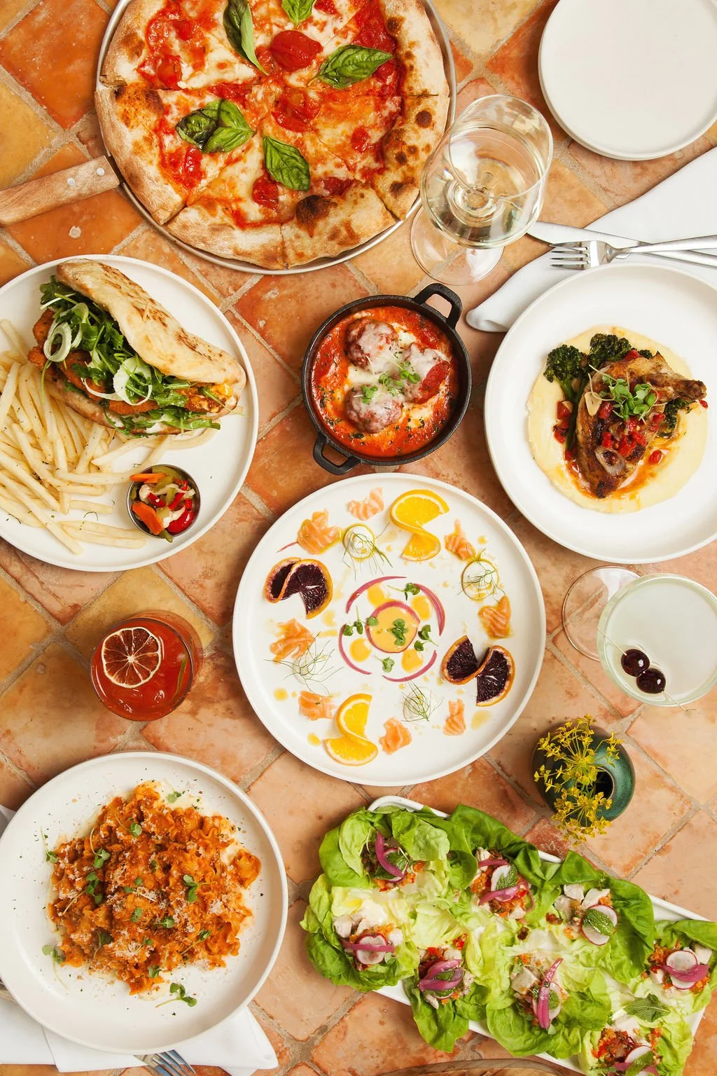 A table with assorted Italian dishes, including pizza with basil, pasta with tomato sauce, a meat dish with vegetables, a seafood dish, a salad with lettuce and onions, and several cocktails and drinks.