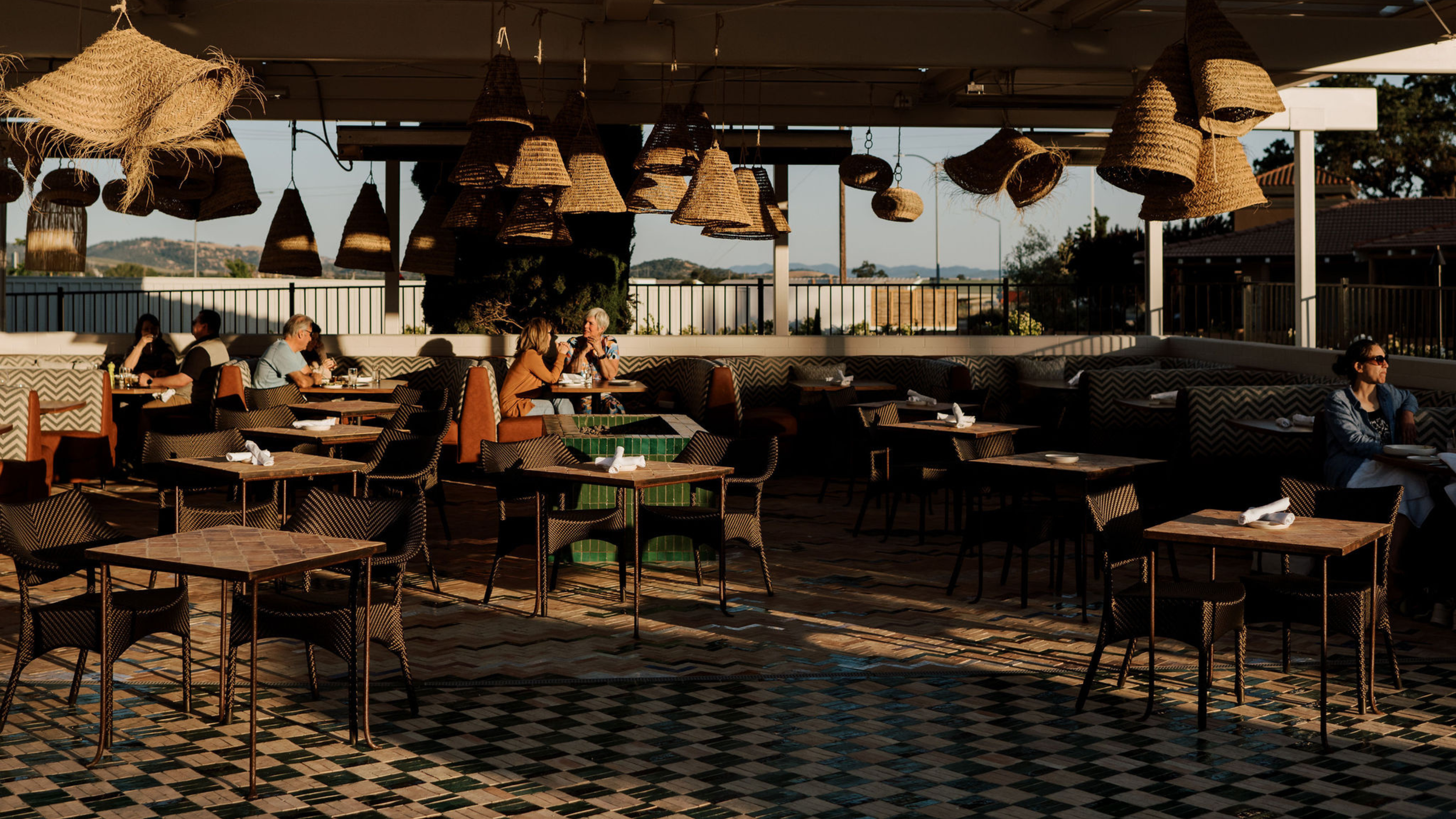 An outdoor restaurant patio with several tables and chairs, hanging woven lamps, and people dining and conversing in the late afternoon sunlight. The area is surrounded by a railing and scenic views of hills and trees in the background.